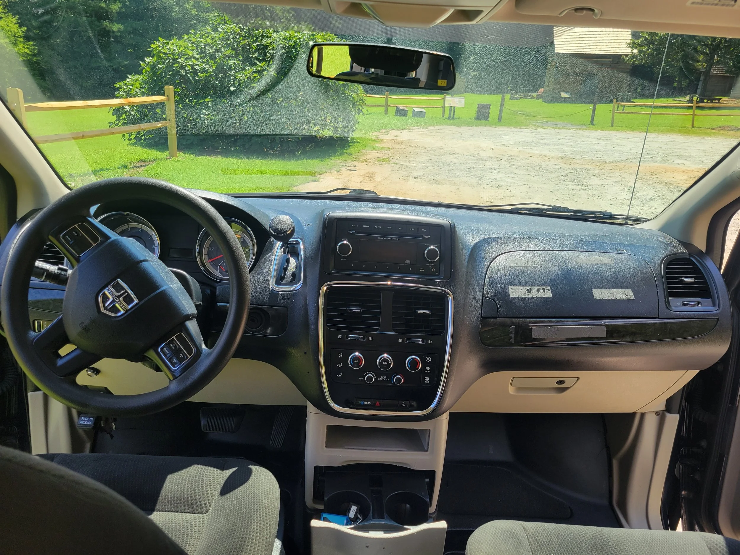 Inside view of a vehicle's dashboard looking through the windshield at a dirt road with green grass, bushes, and trees, and a wooden fence on the roadside.