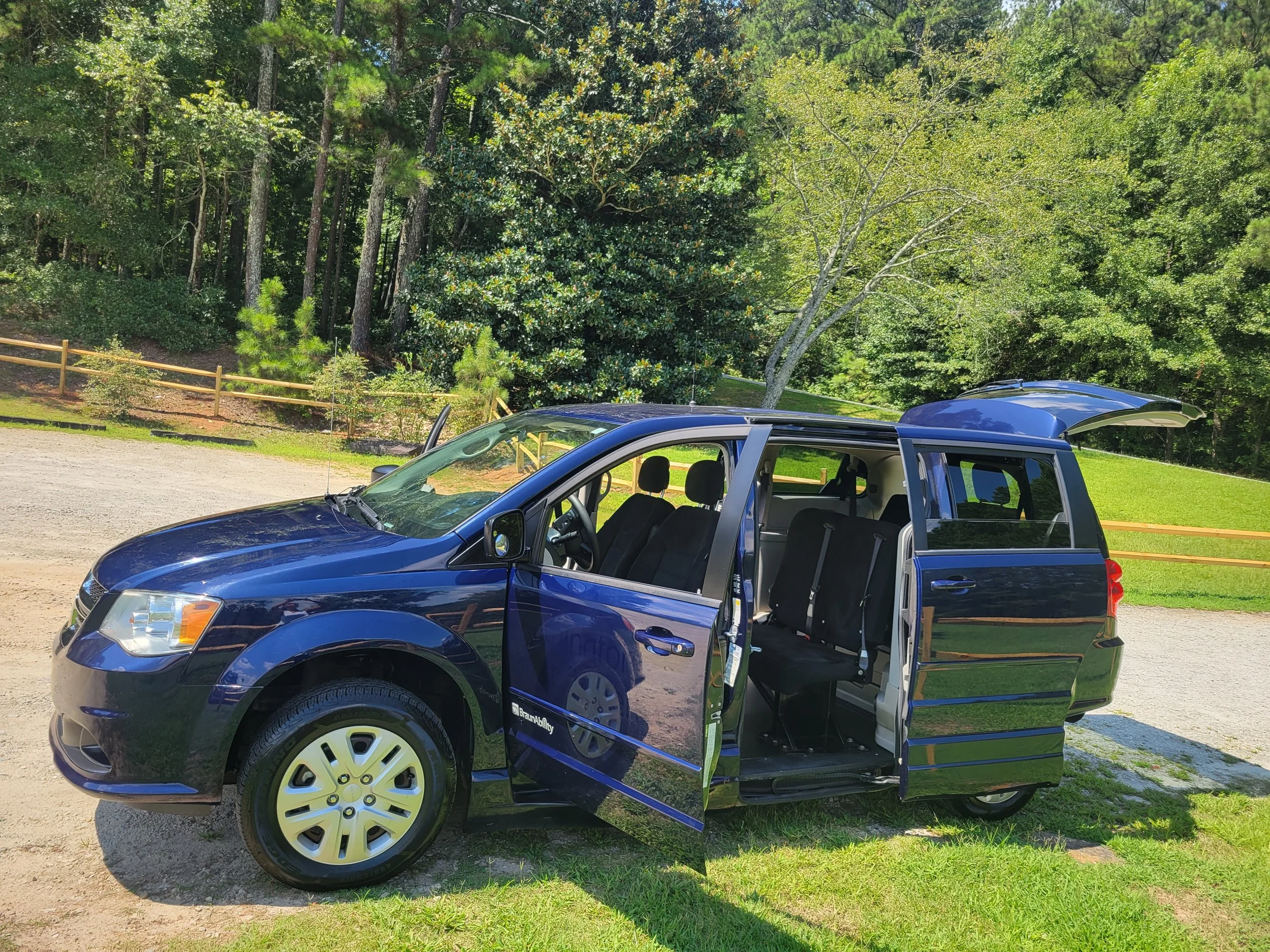 A blue SUV with an open rear passenger door parked on a gravel lot near a wooded area with green trees and grass.