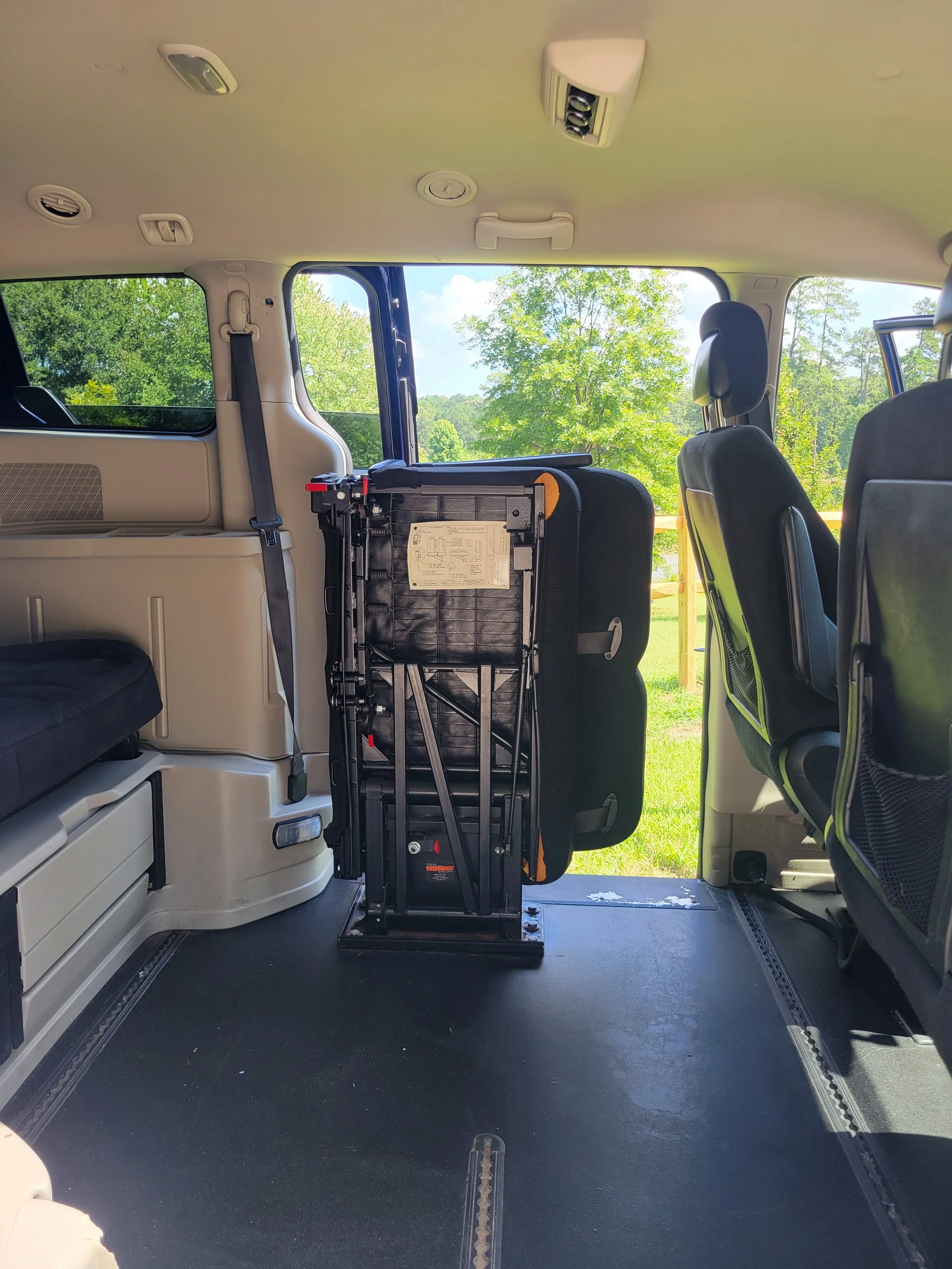 The interior of a vehicle with seats and a folding wheelchair installed, showing a view of green trees and sky outside through the window.