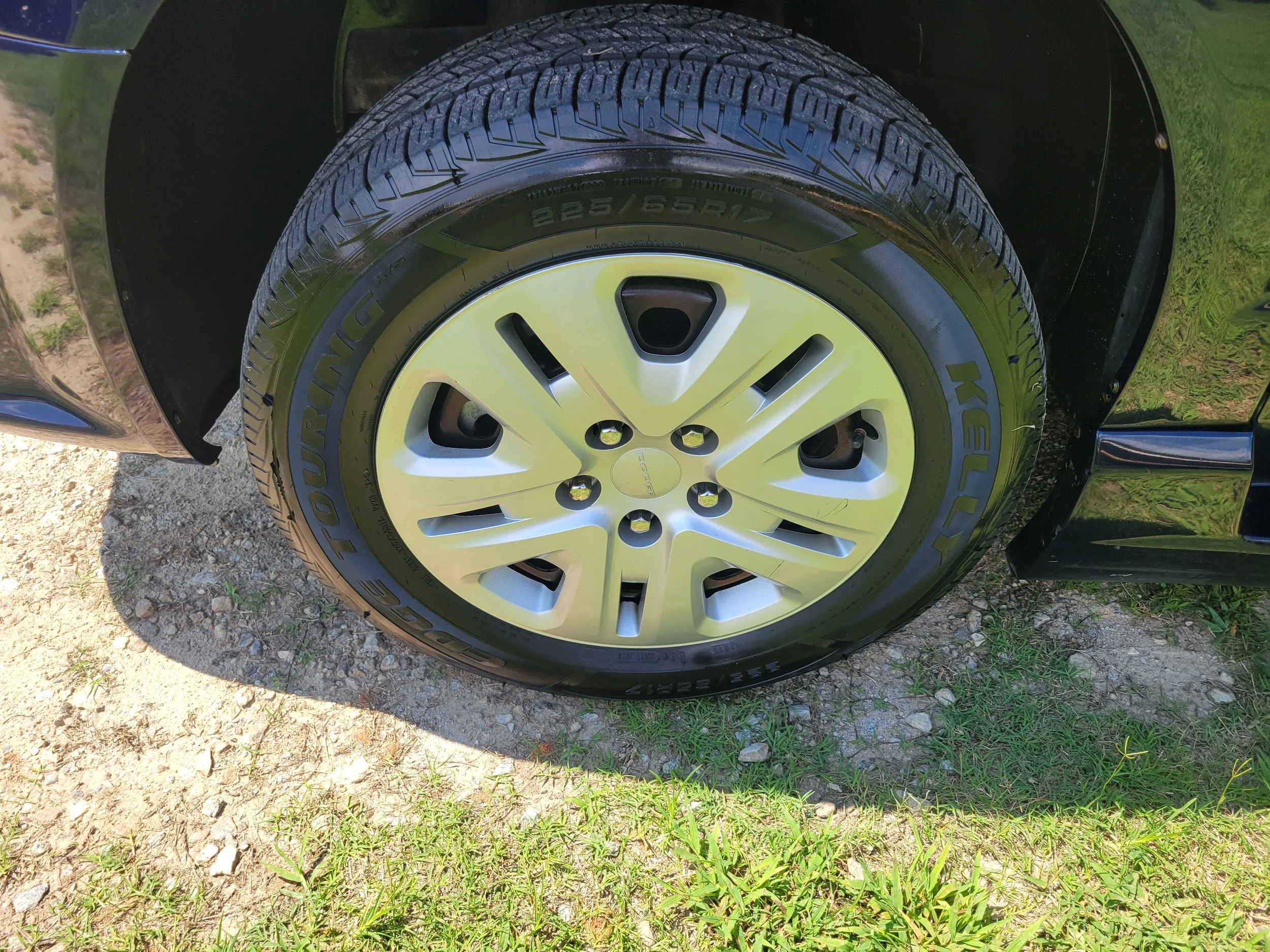 The image shows a close-up view of a car's tire and wheel, with part of the black body and a grassy area with dirt and small rocks around the tire.