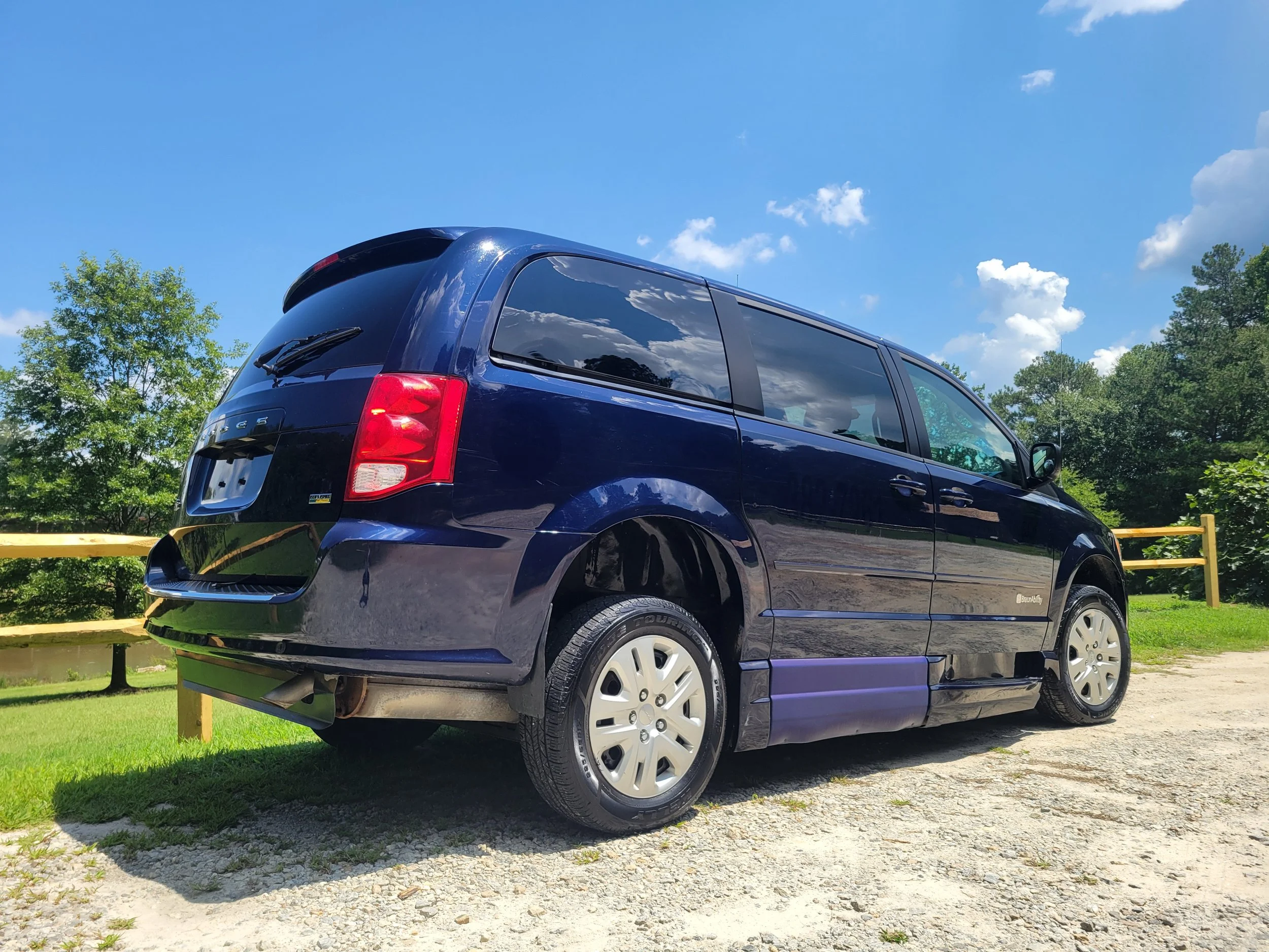Blue minivan parked on a gravel lot with trees and a blue sky in the background.