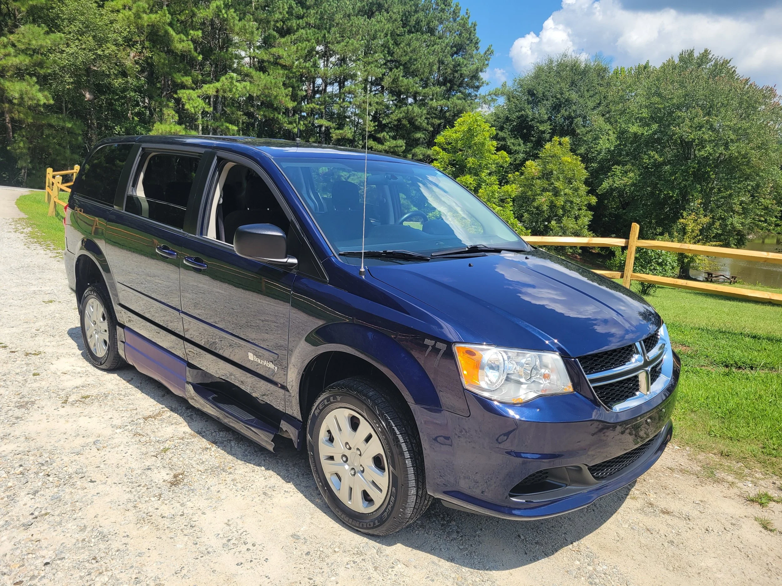 A blue minivan parked on a gravel area next to a green grassy field with trees in the background and a wooden fence, under a partly cloudy sky.