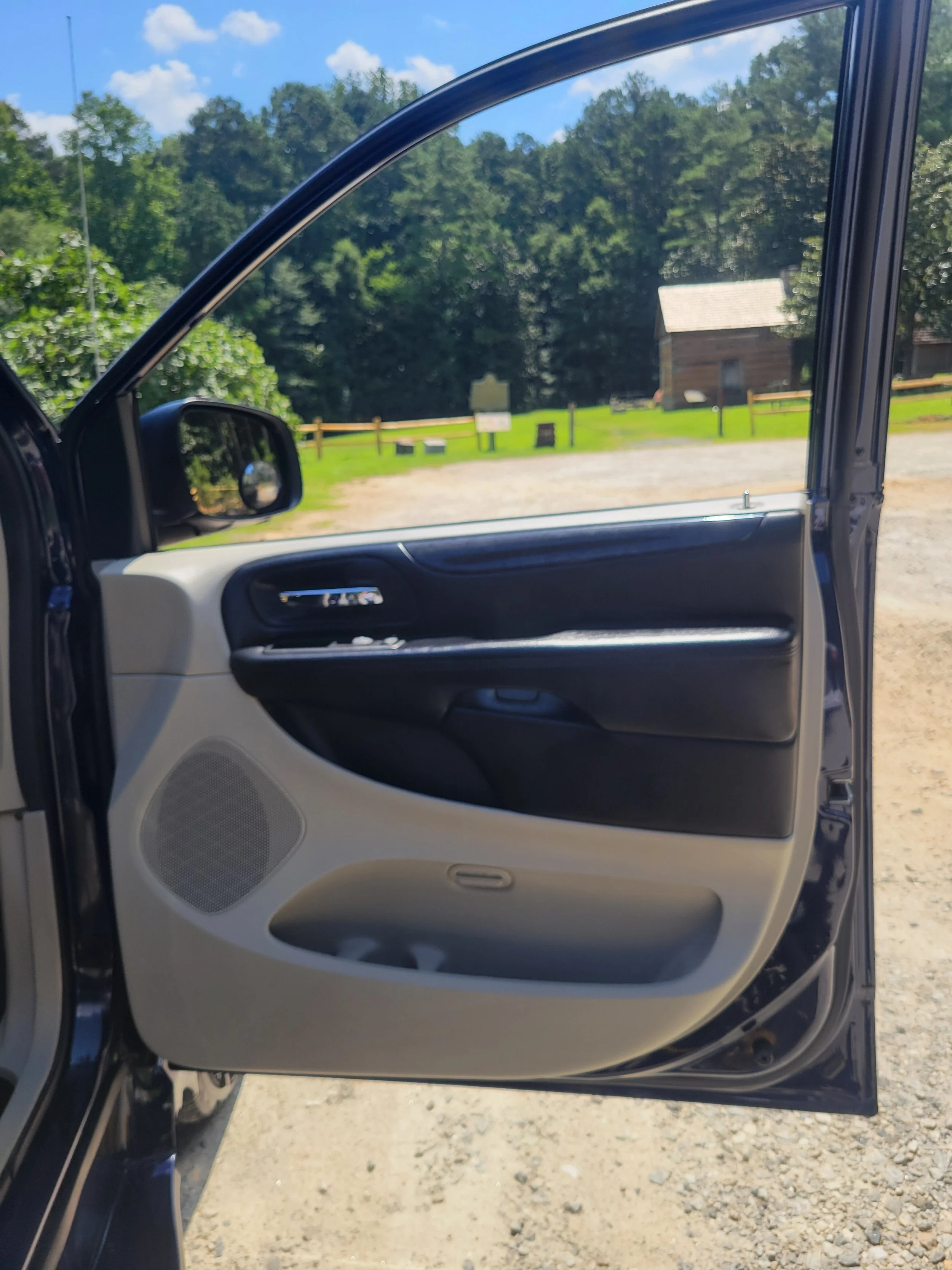 Open car door showing the interior, with a rural background of trees, a small wooden building, and a bright blue sky with clouds.