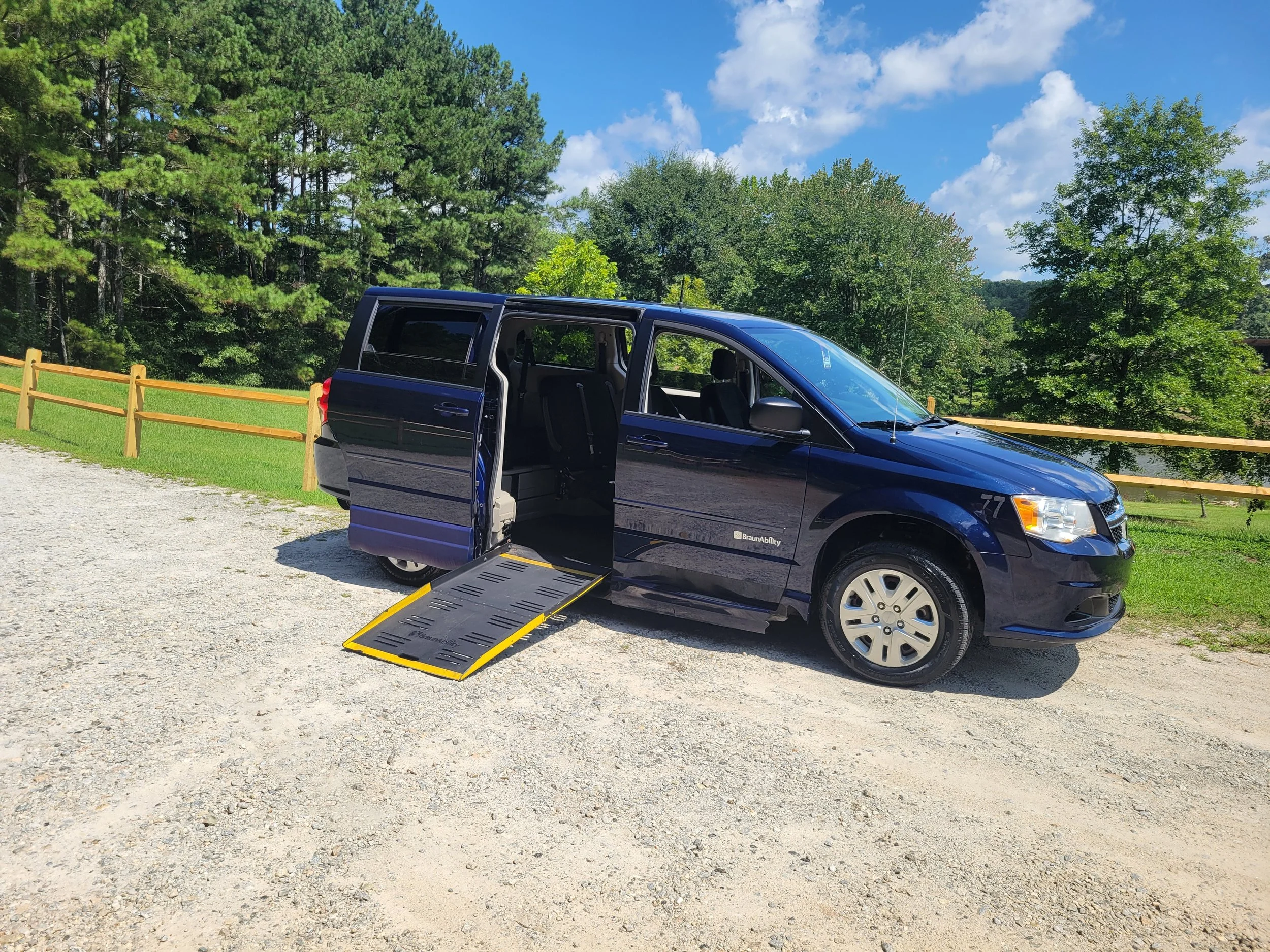 A dark blue minivan with a wheelchair ramp extended, parked on a gravel area with a yellow wooden fence and green trees in the background under a partly cloudy sky.