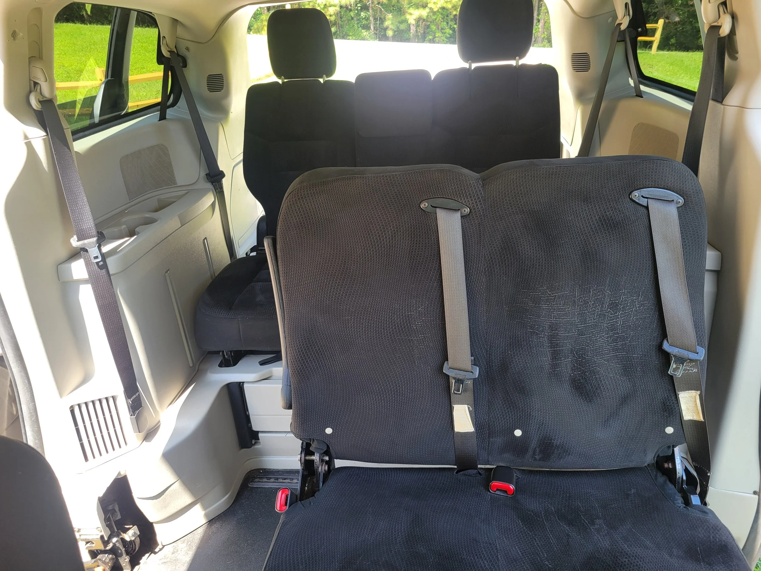 Interior view of a vehicle showing two rows of black fabric seats with seat belts, and side windows revealing greenery outside.