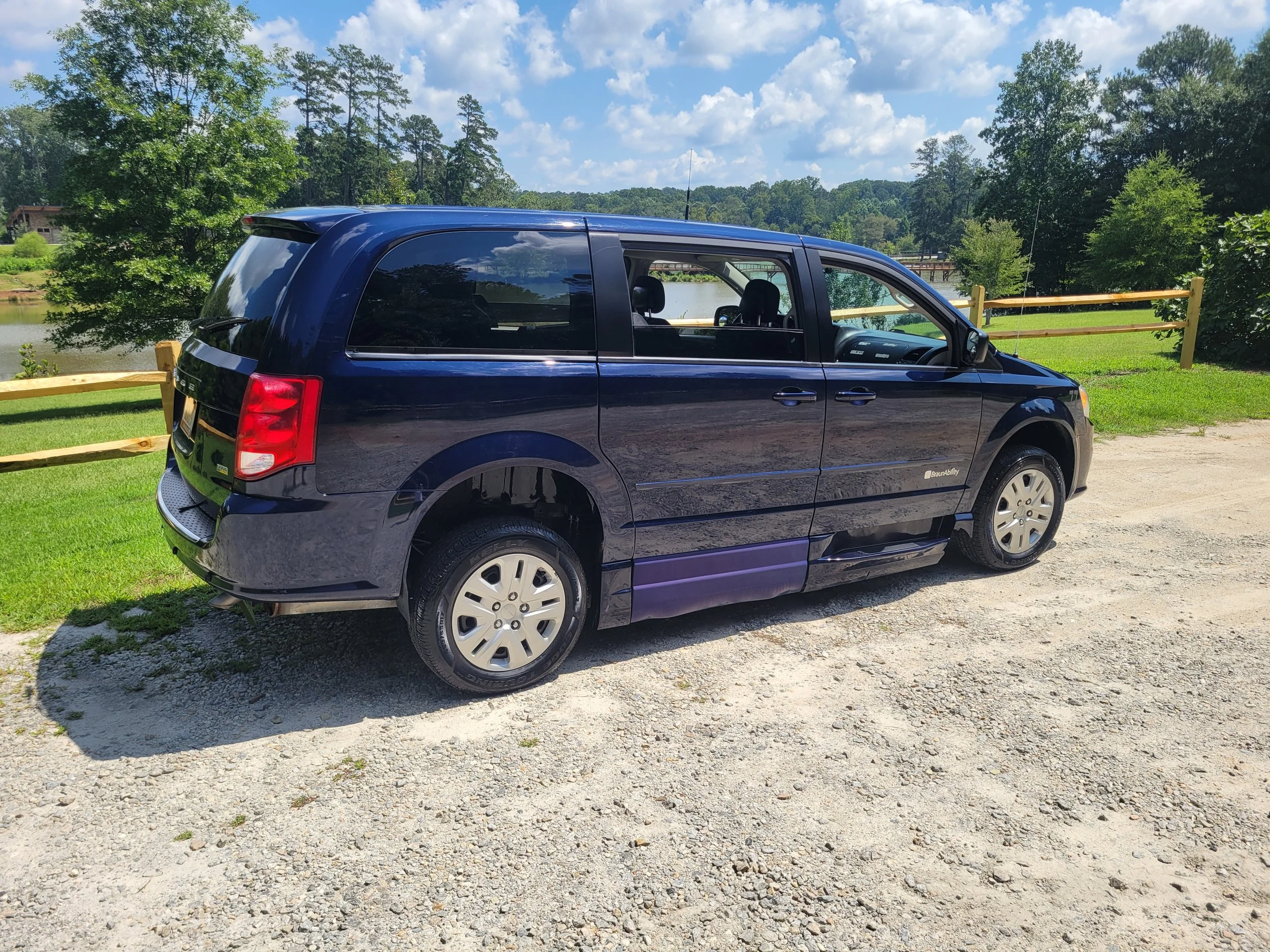 A black minivan is parked on a gravel area near a grassy field with trees and a body of water in the background, under a partly cloudy sky.