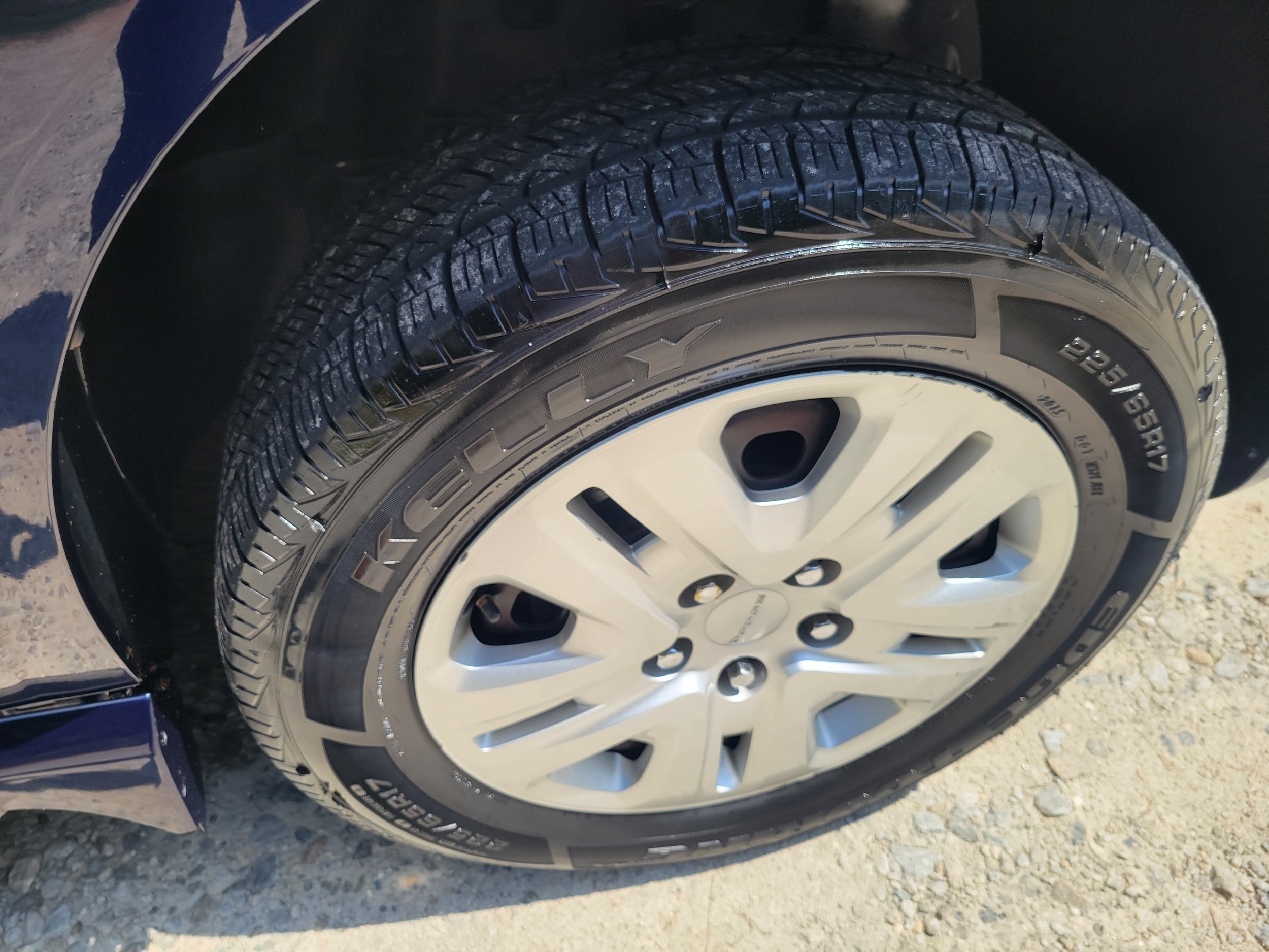 Close-up of a car tire with a detailed tread pattern, mounted on a silver wheel, on a gravel surface.