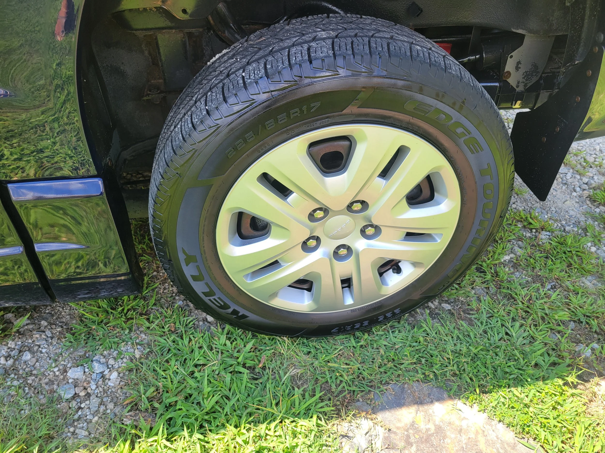 A close-up of a black vehicle wheel with a silver rim and tire, parked on grass and gravel, with the vehicle's fender, suspension, and part of the body visible.