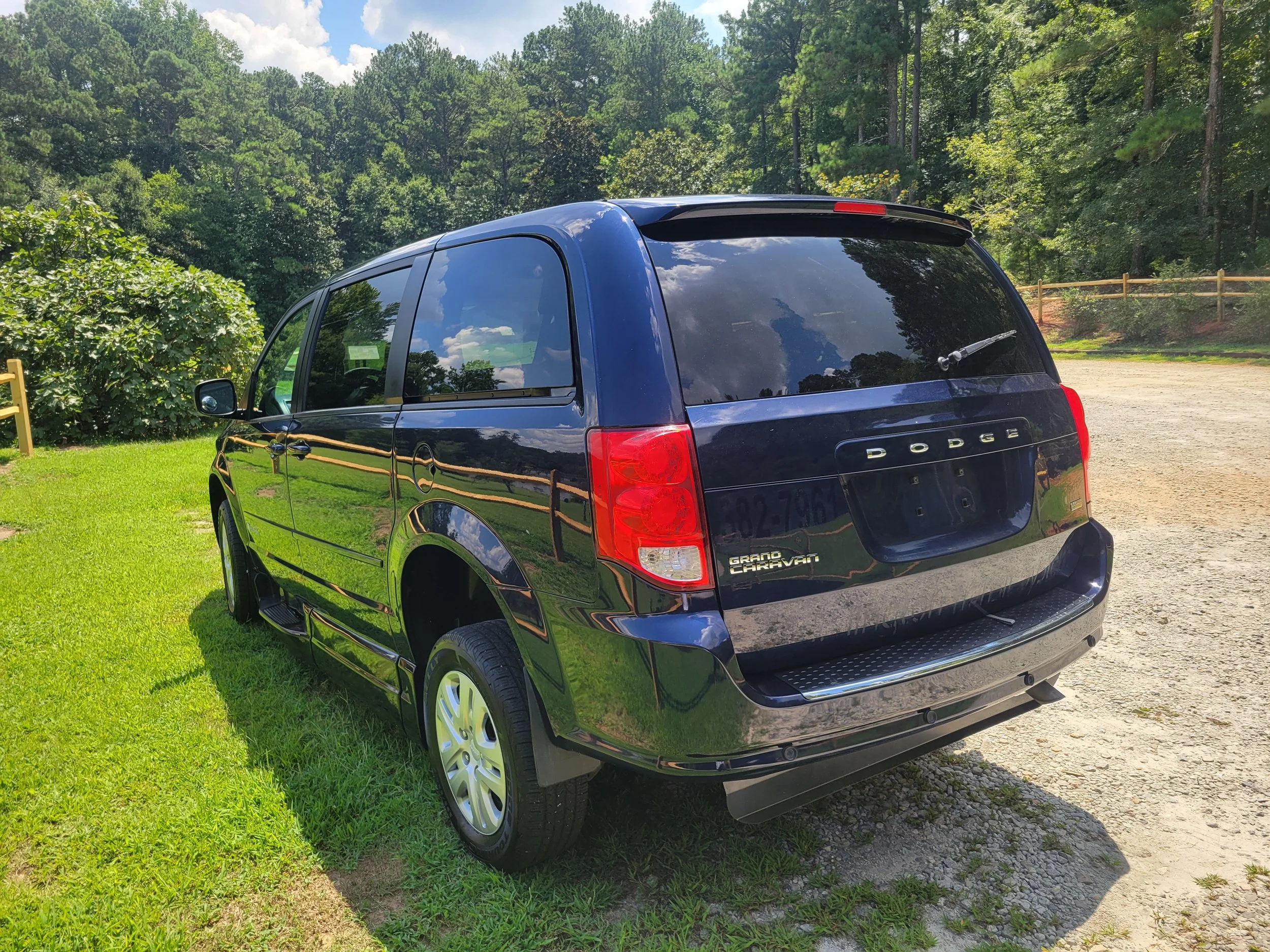 A black Dodge Grand Caravan minivan parked on grassy ground with trees and a wooden fence in the background under a partly cloudy sky.