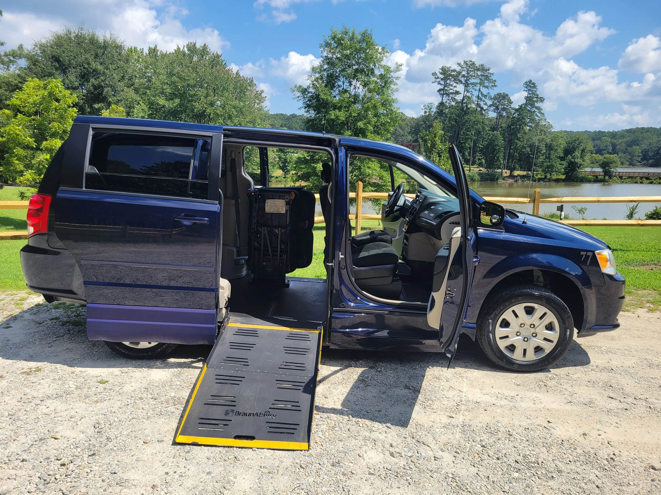 A blue van with the sliding door open and a wheelchair ramp extended by a lake surrounded by green trees and a wooden fence.