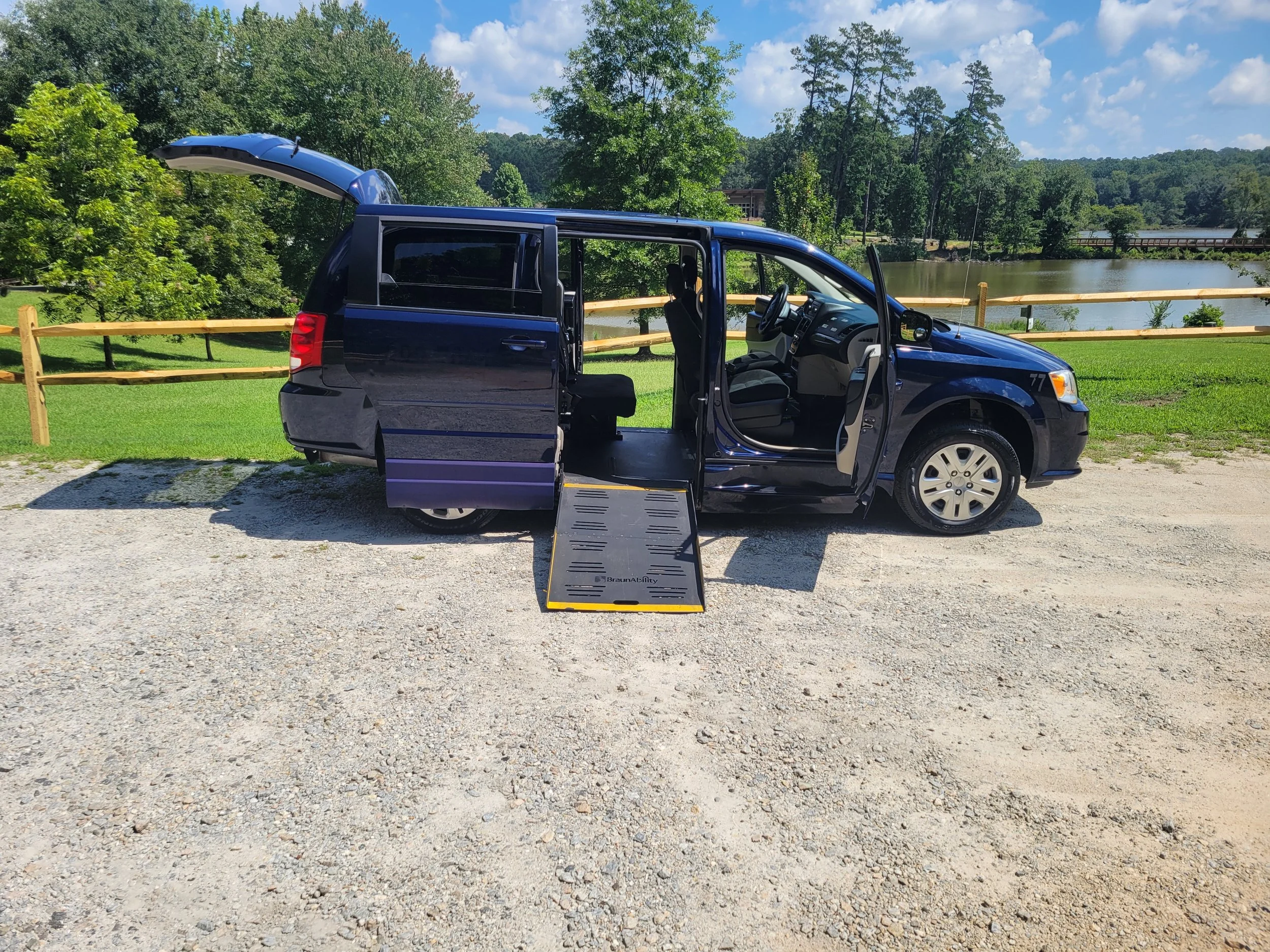 A blue van with a ramp extended to the ground, parked on gravel near a wooden fence, with green trees, a body of water, and a partly cloudy sky in the background.