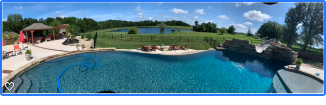 Residential backyard with a swimming pool, poolside lounge chairs, and a covered patio area with outdoor seating, overlooking a lush green field and a pond with trees in the background.