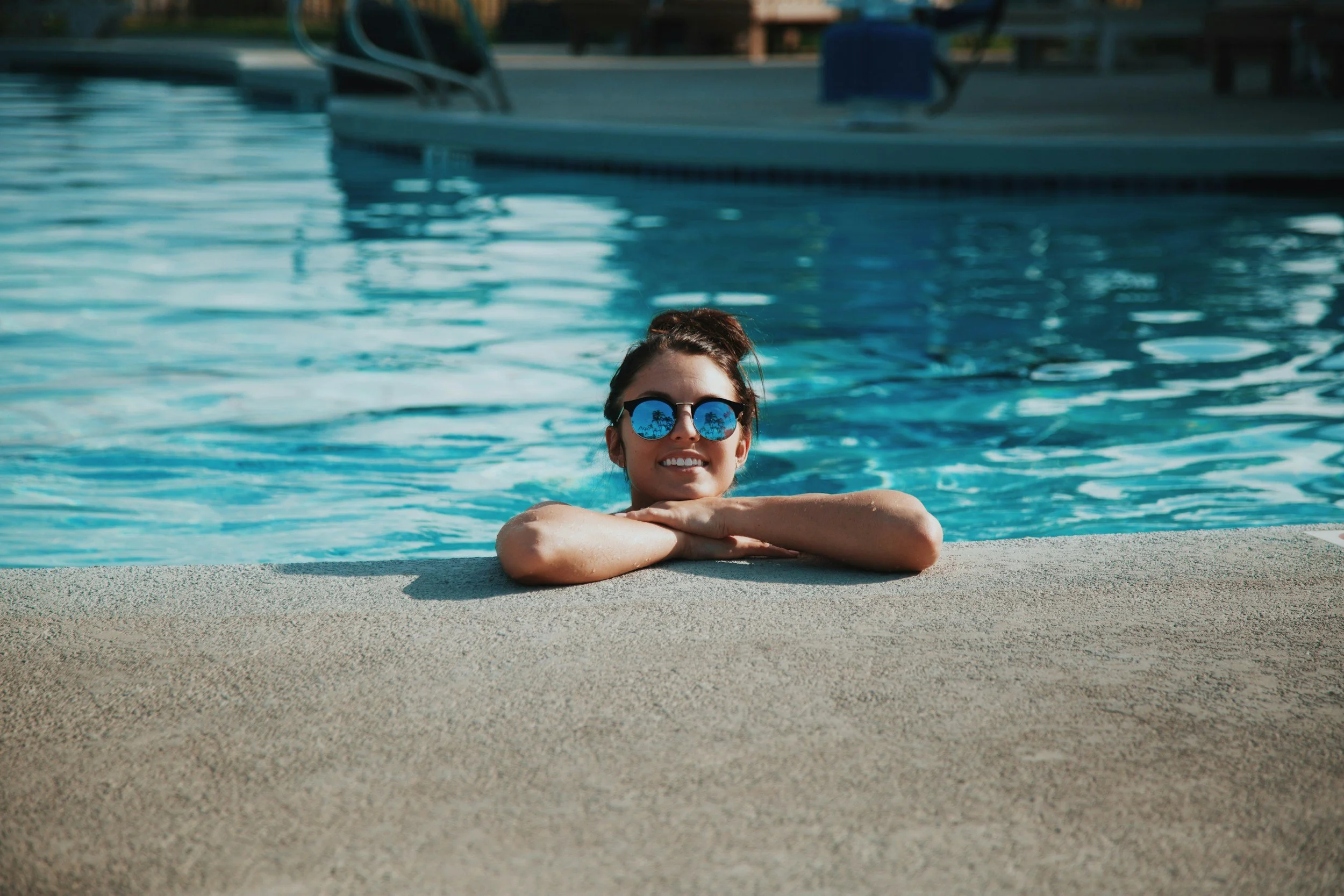 A woman with sunglasses leaning on the edge of a swimming pool, smiling.