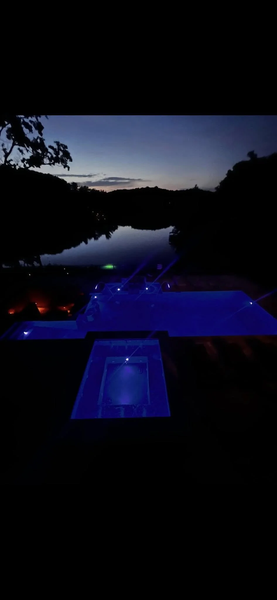 Nighttime view of a dockside area with a swimming pool illuminated by blue lights, overlooking a calm river with a sunset sky in the distance and tree silhouettes on the horizon.
