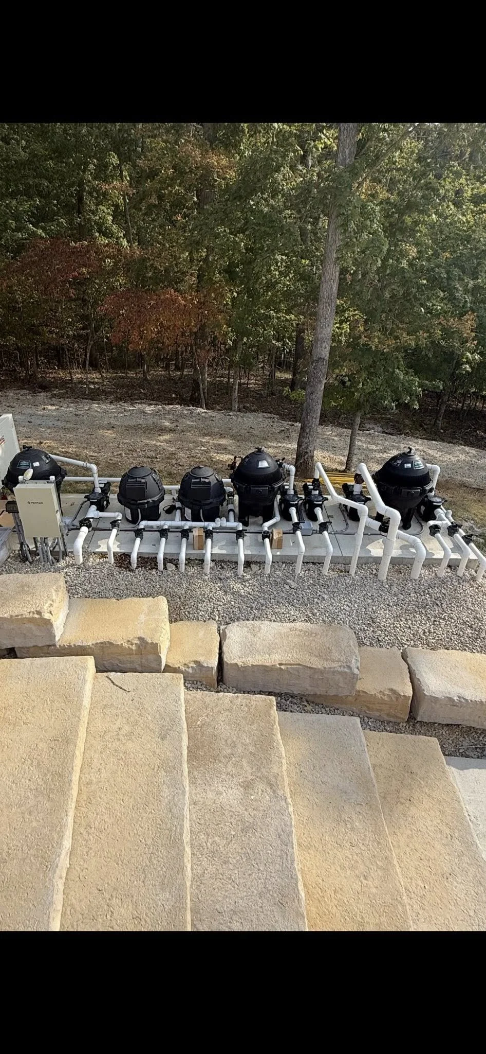 Outdoor pool pump system with black circular filters, white pipes, and valves, installed on a concrete slab behind a stone patio with yellowish stone steps, surrounded by a natural landscape with trees and earthy ground.