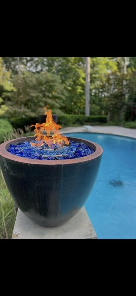 An outdoor scene with a black pot containing blue glass and orange flames, positioned near a swimming pool surrounded by green trees and shrubs.