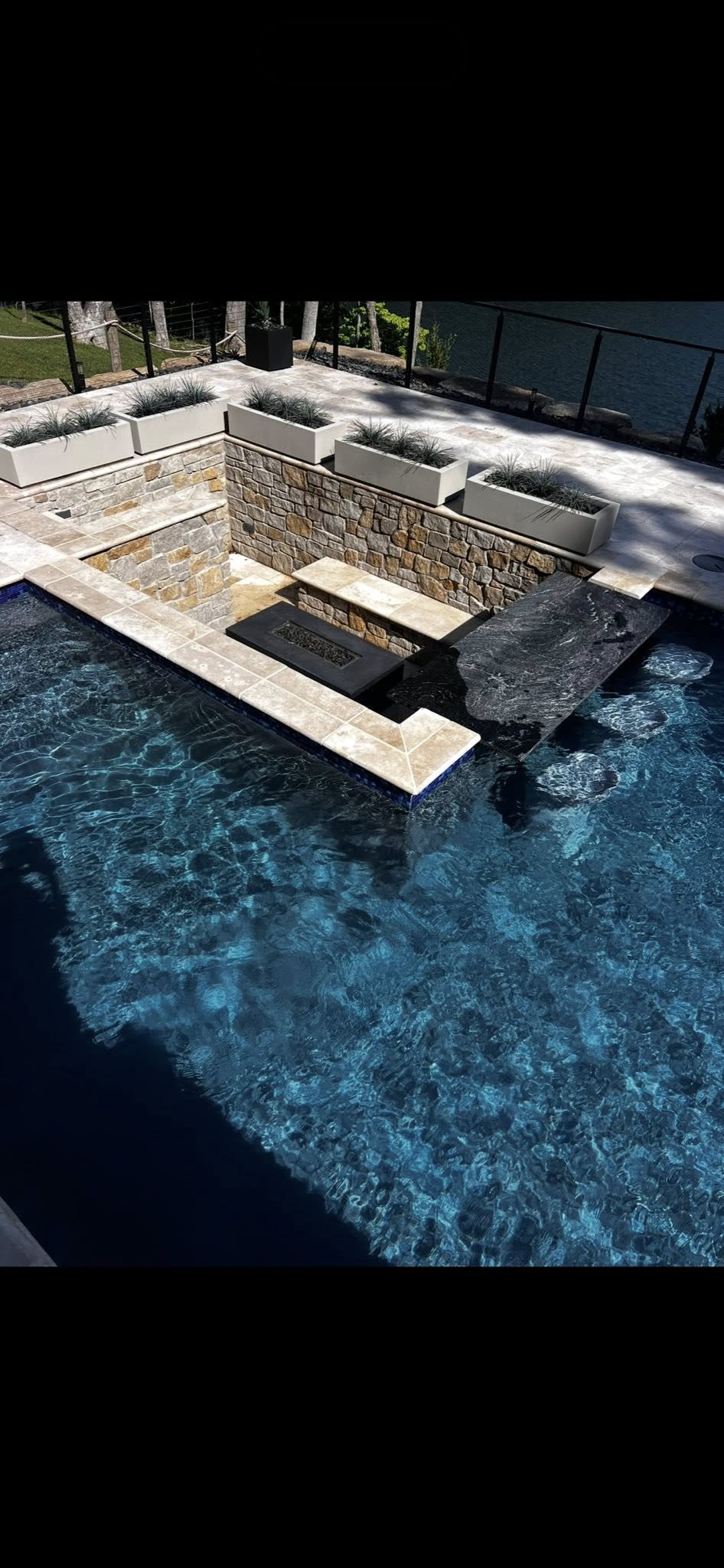A swimming pool with a stone waterfall feature, surrounded by tiled decking and potted plants, overlooking a body of water and trees in the background.