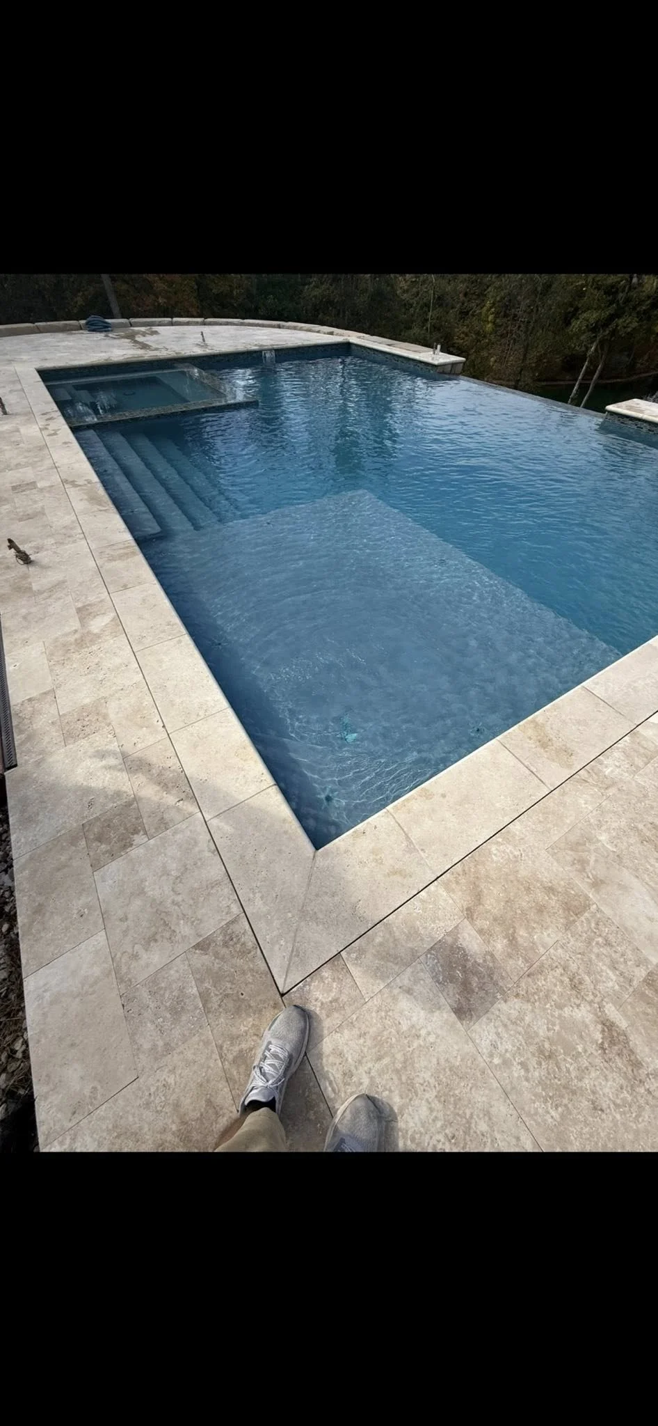 A rectangular swimming pool with steps at one end, surrounded by beige stone tiles, with a person's sneakers visible in the foreground.