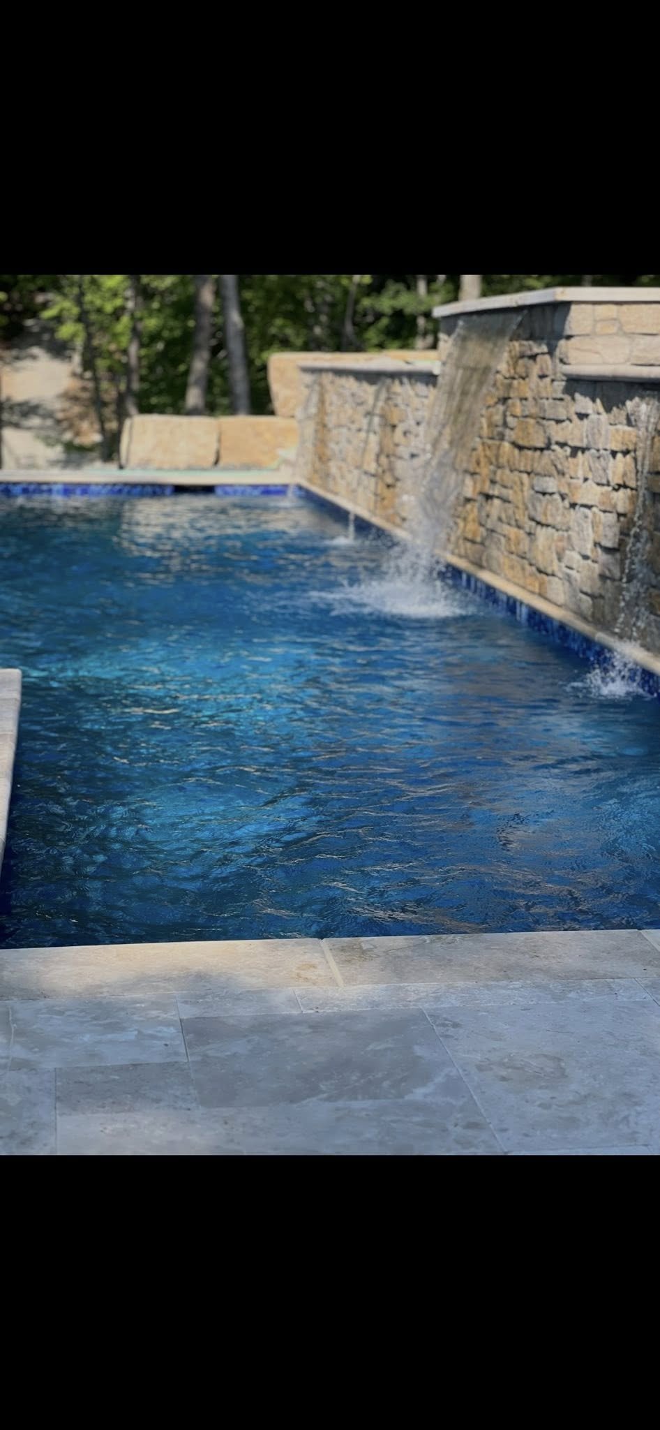 View of a swimming pool with water cascading over a stone wall in the background, surrounded by trees.