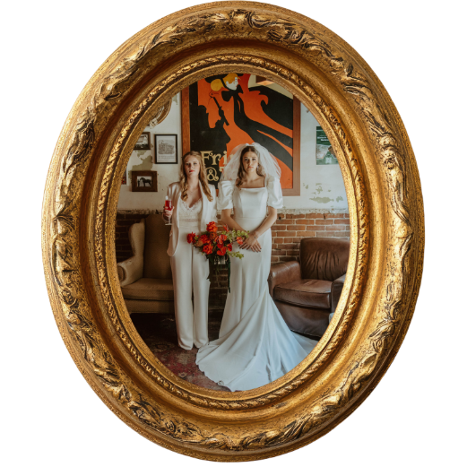 Two women in white wedding dresses standing in front of a mirror with a gold ornate frame, one holding a bouquet of red flowers, in a room with brick wall and artwork.