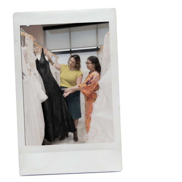 Two women in a gallery of wedding dresses, looking at a black gown in a bridal store