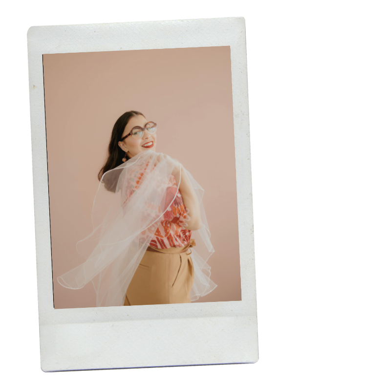 A woman with dark hair and glasses smiling at the camera, wearing a colorful patterned top, tan pants, and holding a veil, against a light pink background.