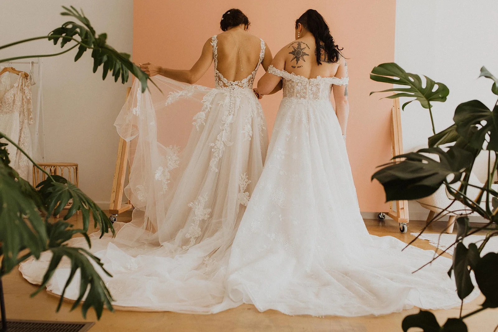 Two women in wedding dresses examining gowns in a room with plants and a pink and white background.