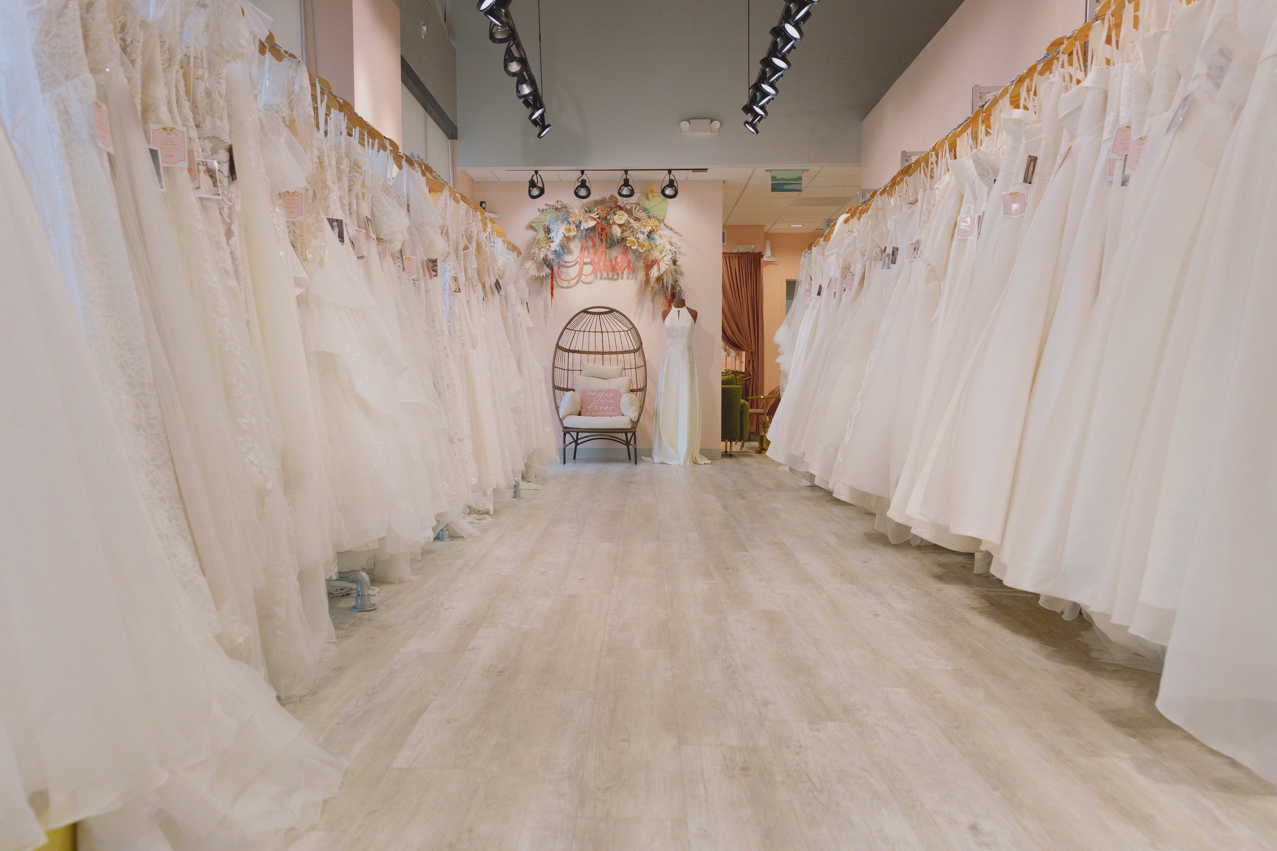 Store aisle with bridal wedding dresses hanging on both sides and a bridal dress mannequin and decorative chair at the back.