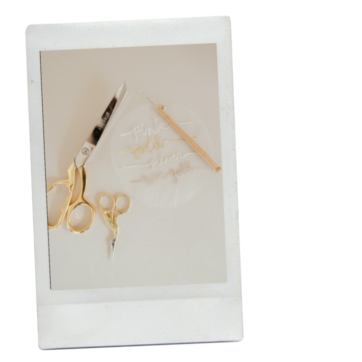 A pair of gold scissors, a pair of small gold scissors, an tambour embroidery hook with gold, silver, and rose gold embroidered writing arranged on a circle of tulle