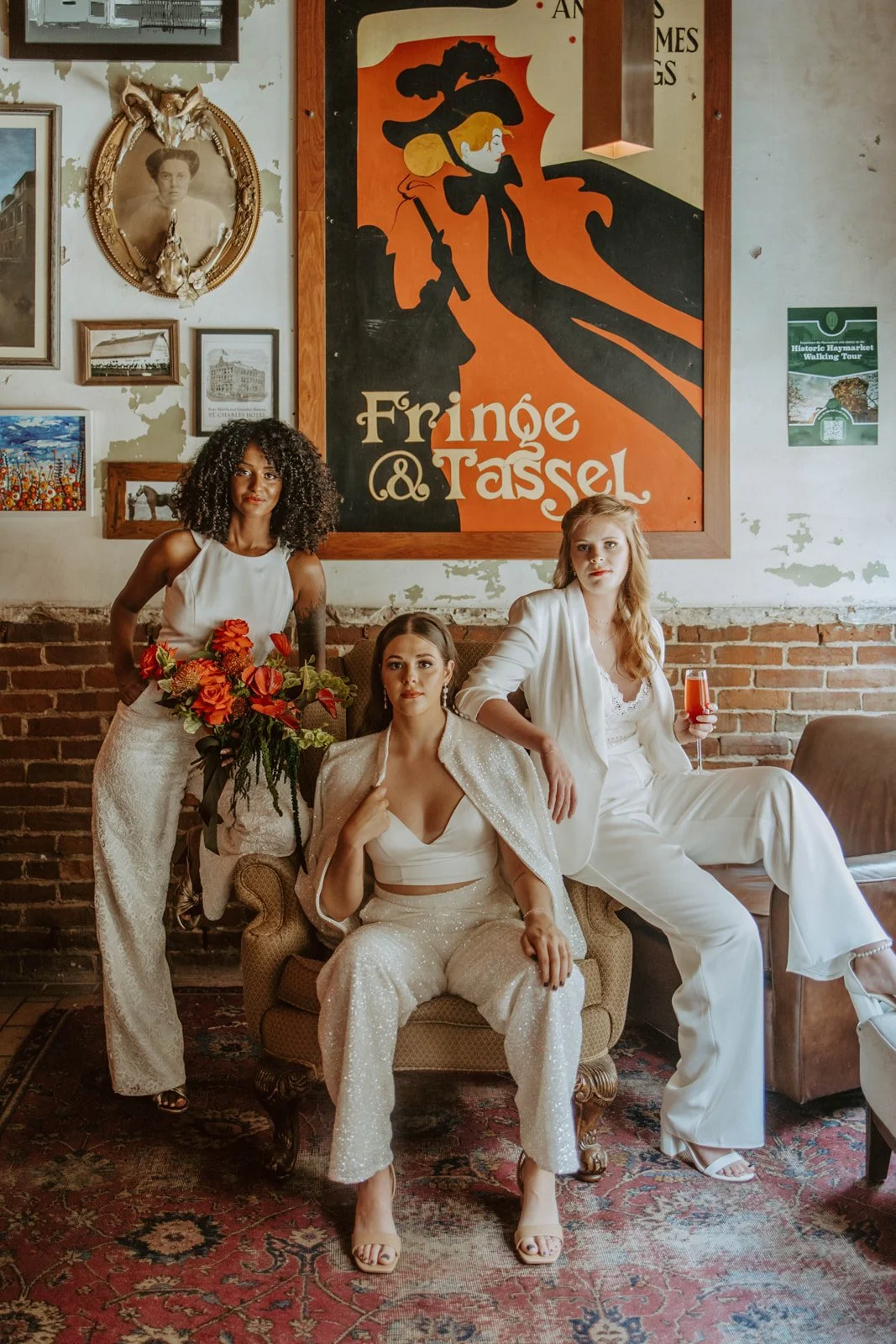 Three women pose inside a vintage-style room with exposed brick walls. The woman in the center is seated on an armchair wearing a sparkling cream-colored outfit. The woman on the left is standing, holding a bouquet of red flowers, and wearing a white top with cream pants. The woman on the right is sitting on a sofa with a glass of red beverage, dressed in a white pantsuit. Behind them is a large, colorful poster with a stylized woman wearing a hat and the text "Fringe & Tasse." The room is decorated with framed pictures and an oval portrait on the wall.