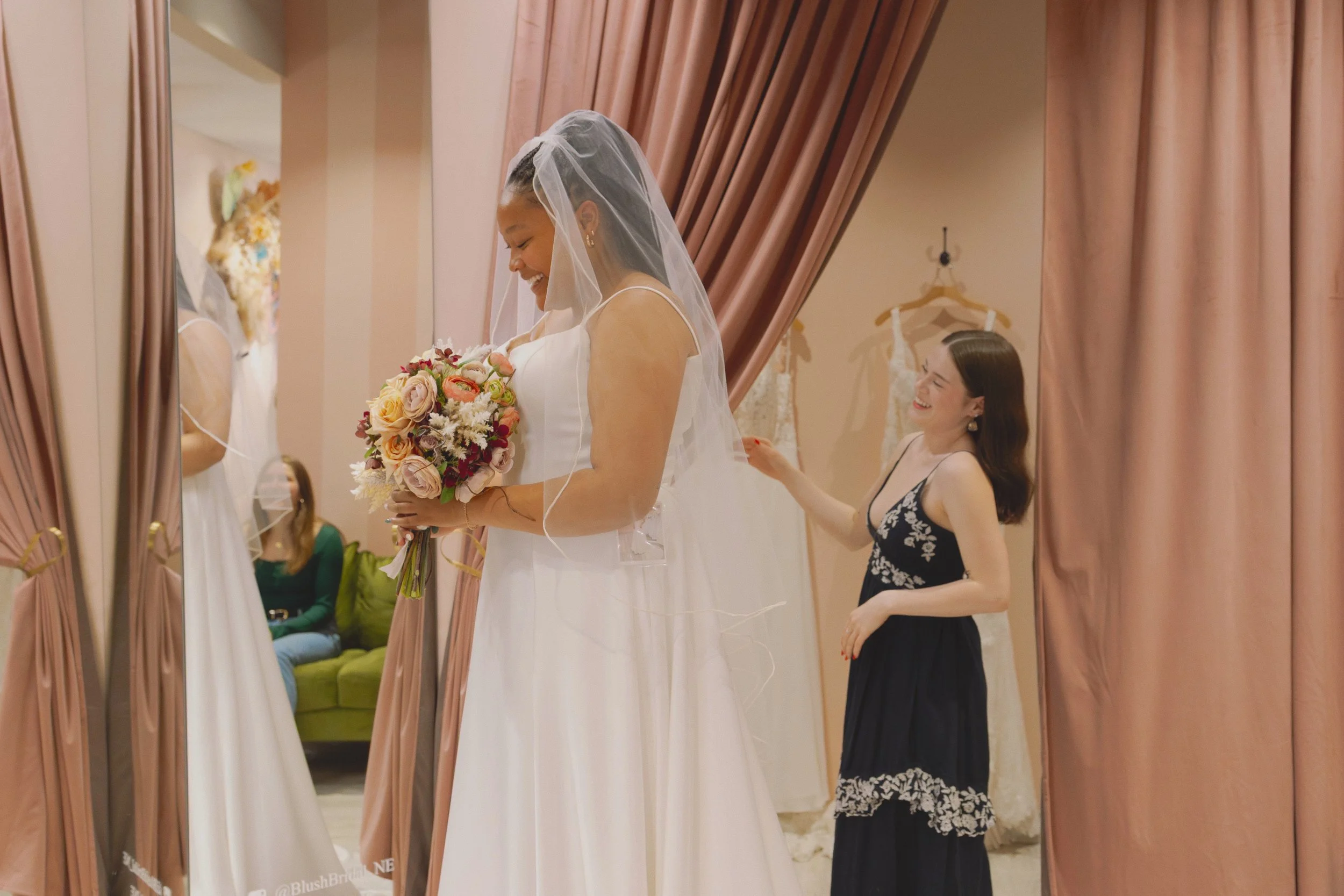 A bride in a white wedding dress holding a bouquet of flowers, smiling. Behind her, a woman in a black dress with white embroidery, laughing and touching her hair. The scene is reflected in a mirror, with pink curtains and wedding dresses hanging in the background.
