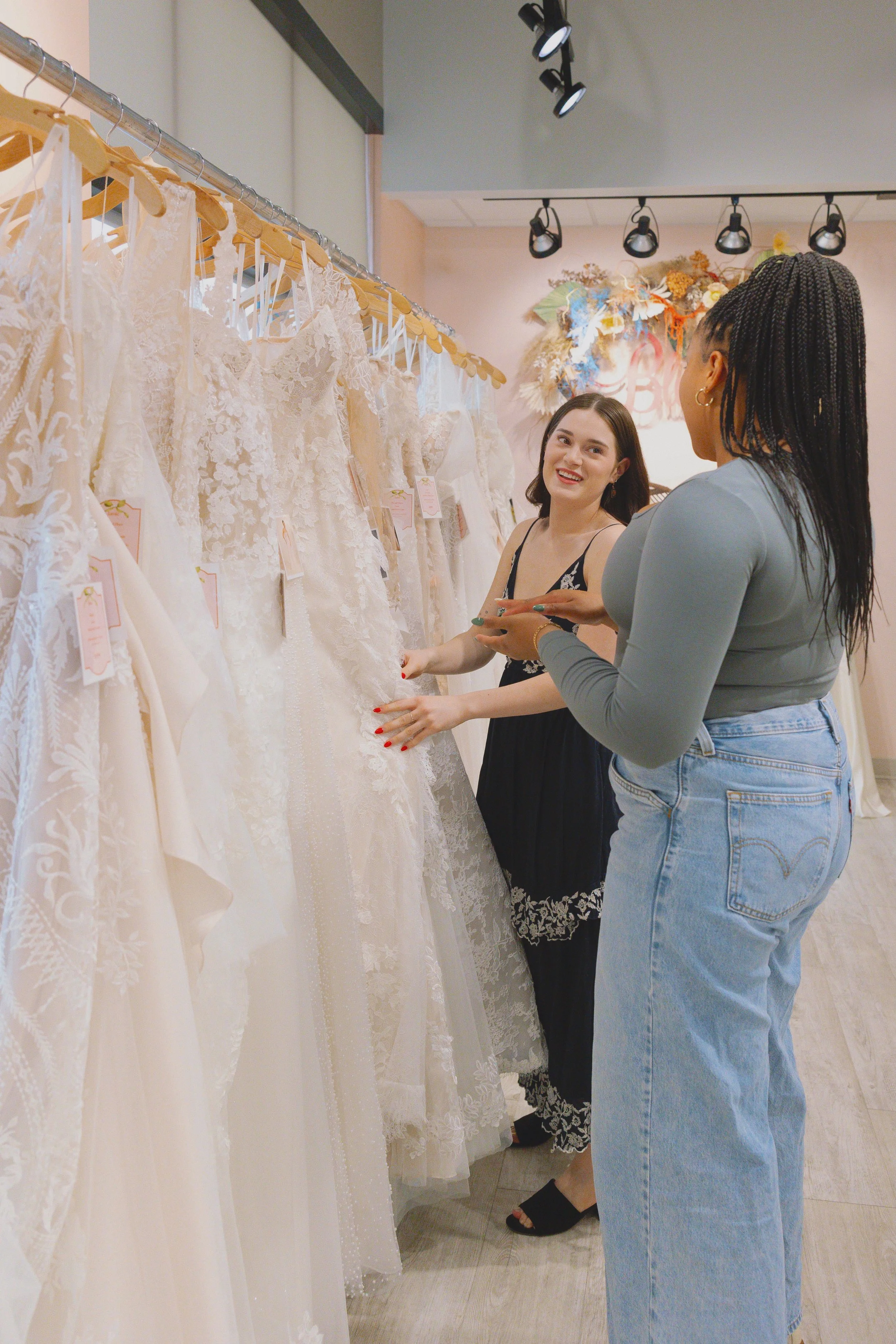 Two women shopping for wedding dresses in a bridal boutique, looking at white lace dresses on a rack, one woman smiling while the other points at a dress.