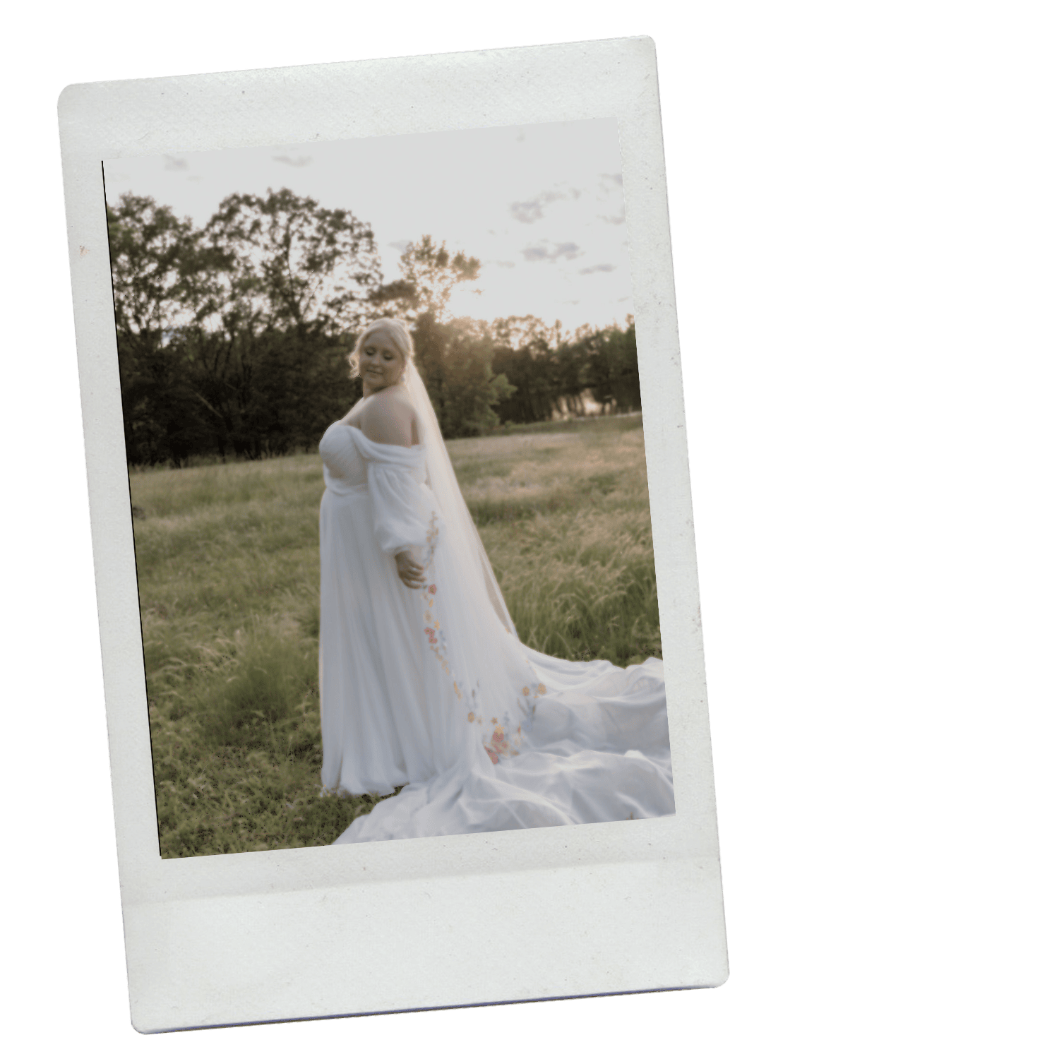 A woman in a white wedding dress with floral embroidery stands in a grassy field at sunset, with trees in the background.