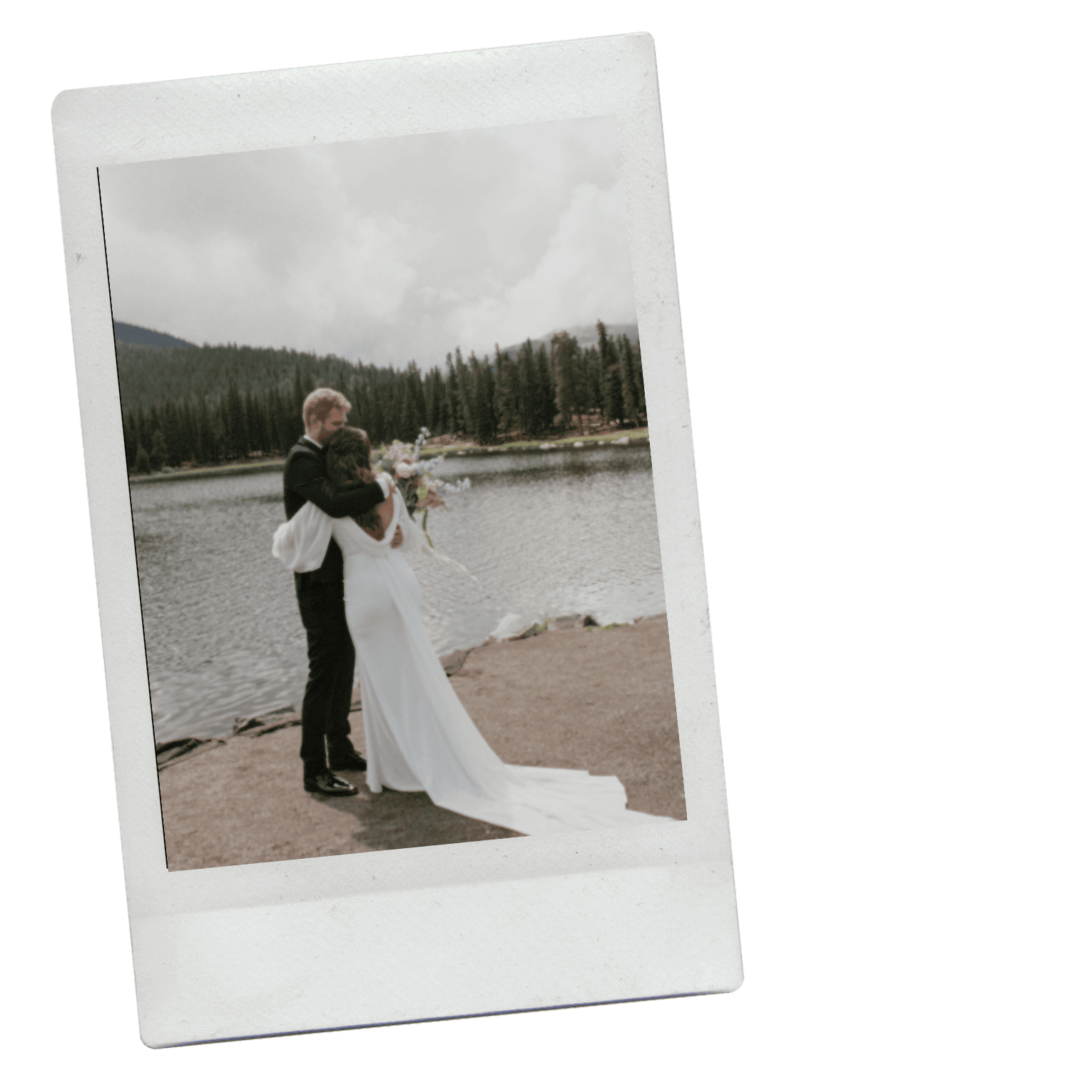 A bride and groom embracing by a lakeside, with trees and mountains in the background, during their wedding day.