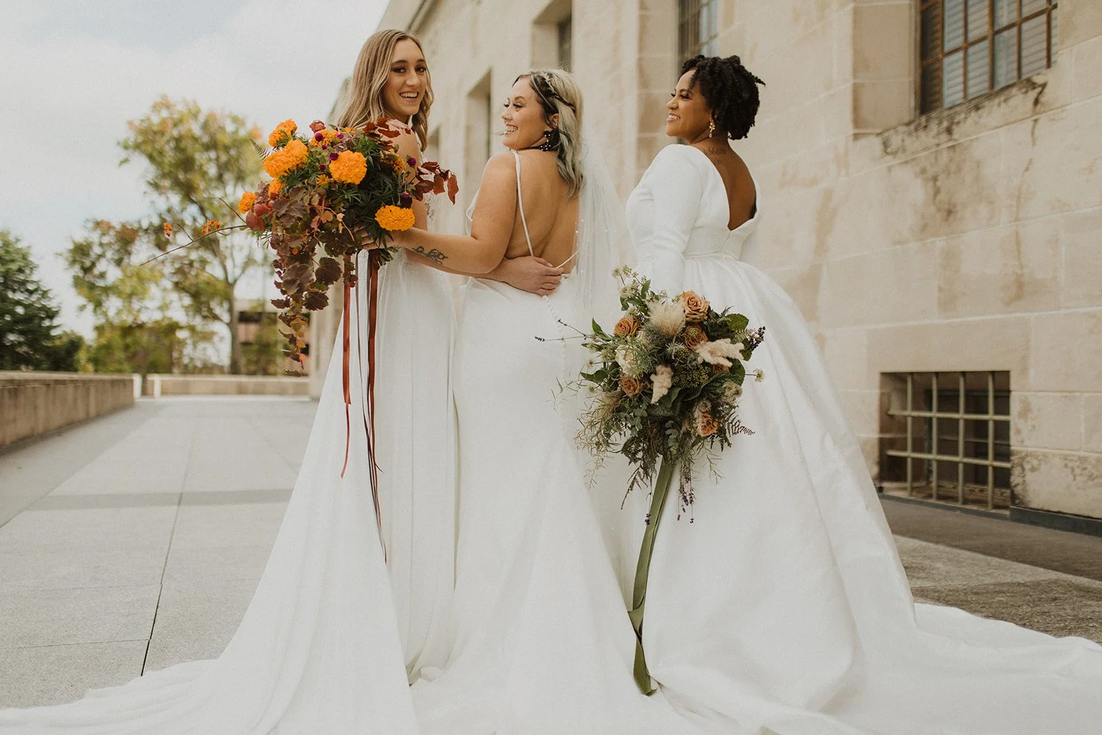Three women in wedding dresses holding bouquets outside a building with a stone wall.