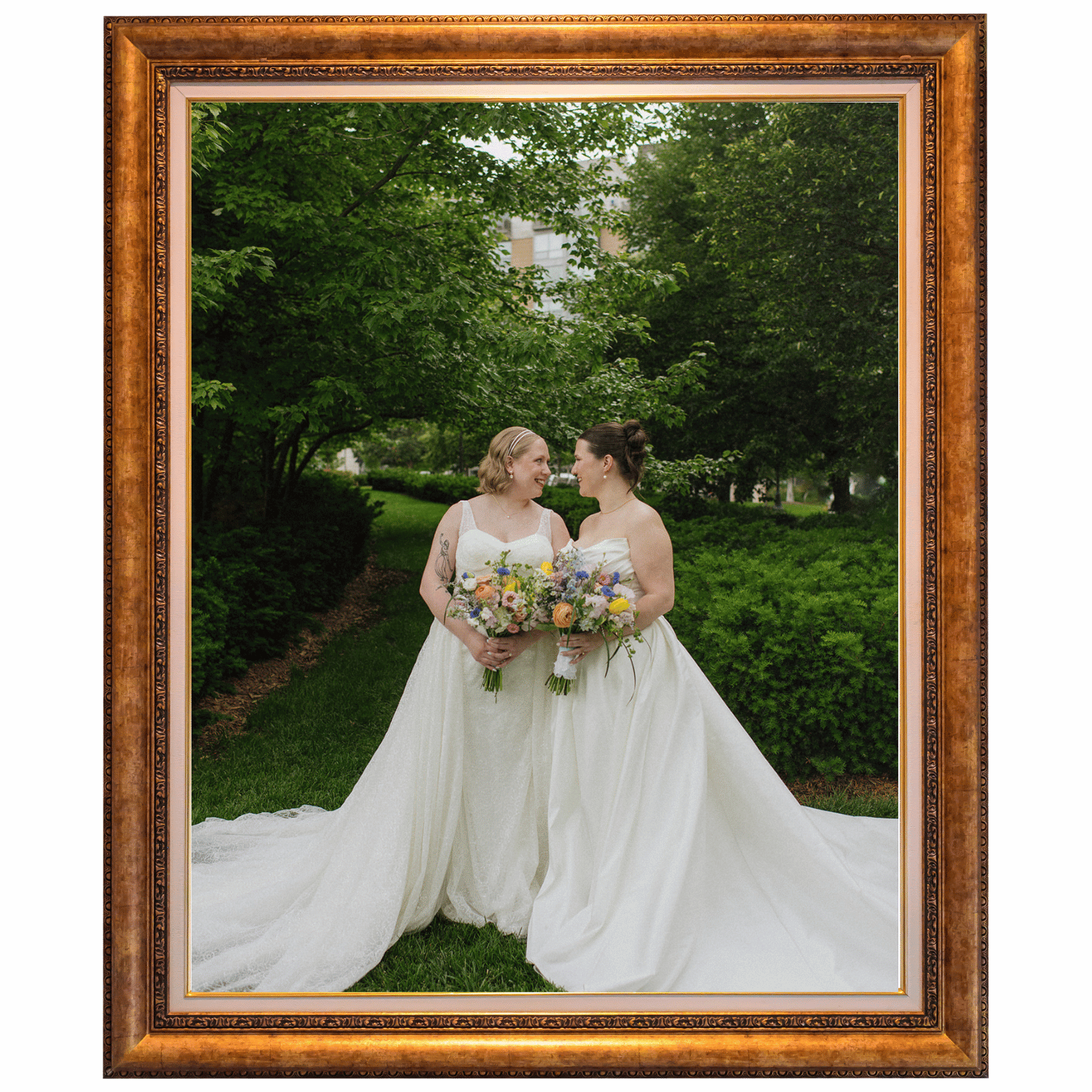 Two women dressed in wedding gowns holding bouquets standing close together in a park surrounded by green trees and bushes, framed in a decorative wooden frame.