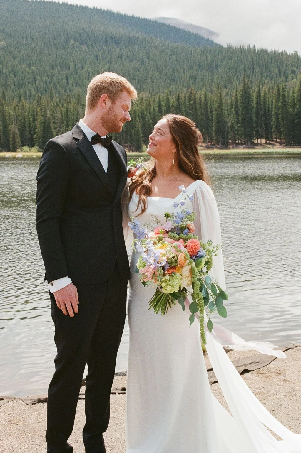 A bride and groom standing by a lake with a mountain and forest background, gazing at each other. The bride holds a colorful bouquet of flowers, and the groom wears a black tuxedo with a bow tie.