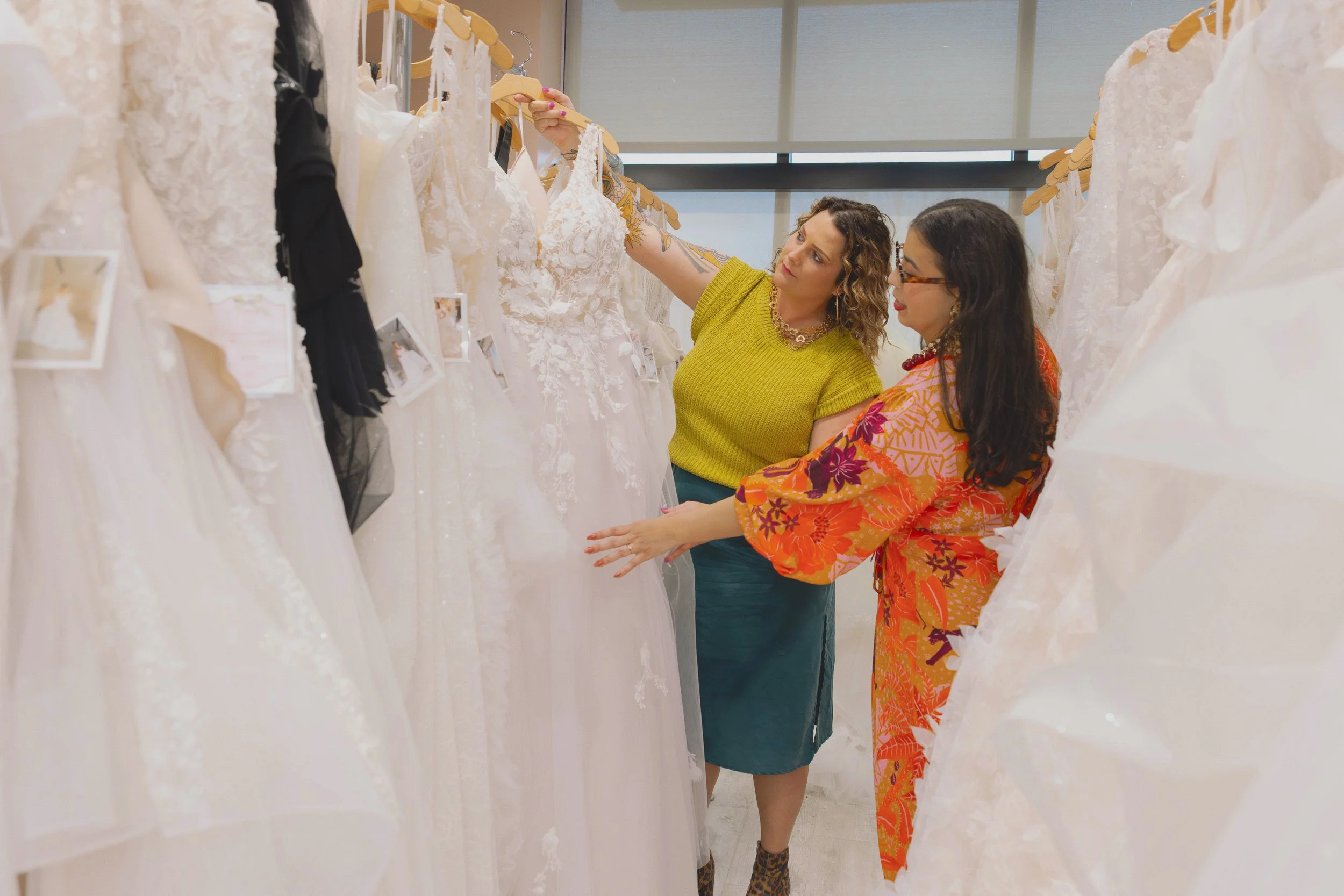 Two women shopping for wedding dresses in a bridal boutique, examining a white wedding gown.