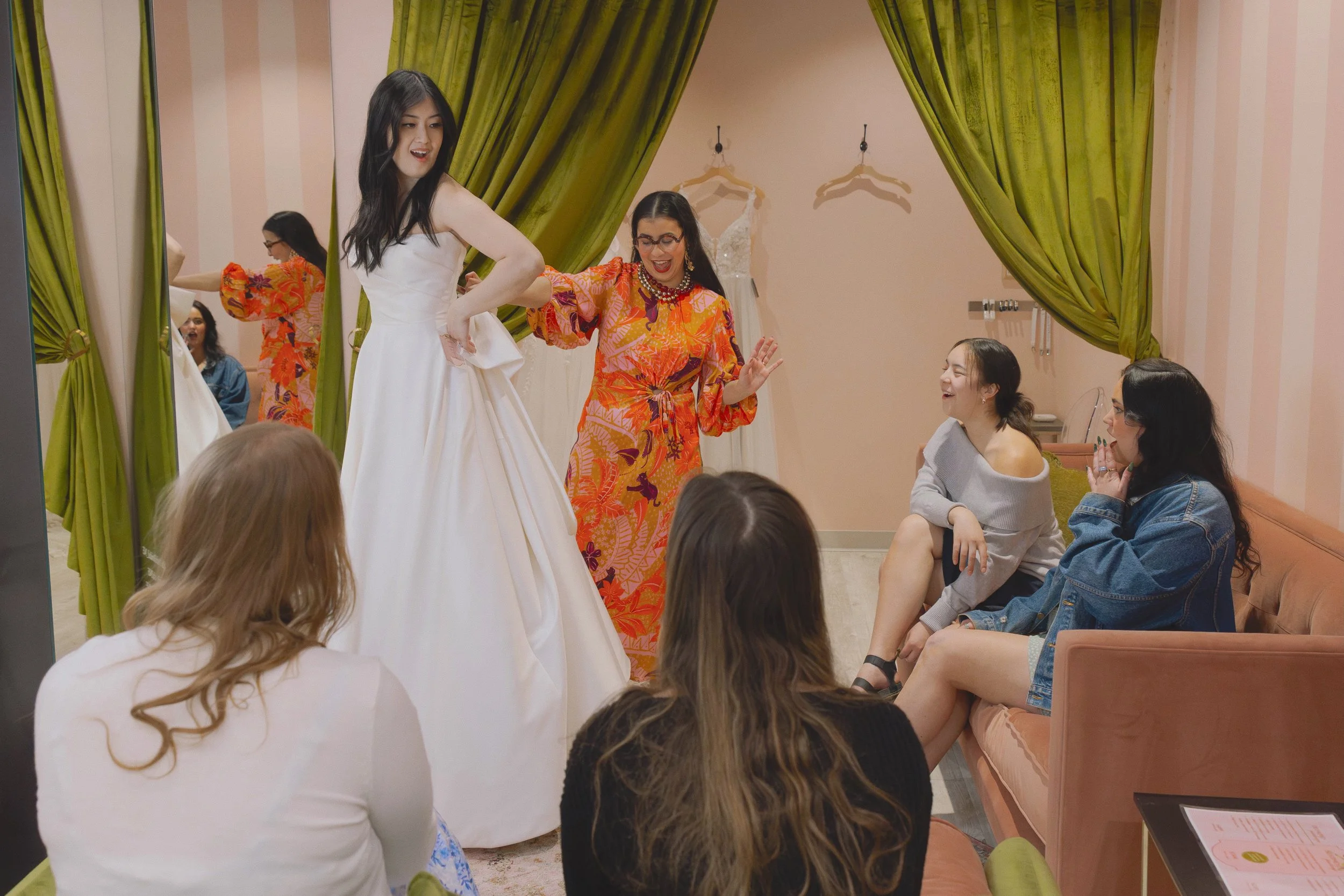 A group of women in a fitting room, one trying on a wedding dress while others watch and cheer, with green curtains and dresses hanging in the background.