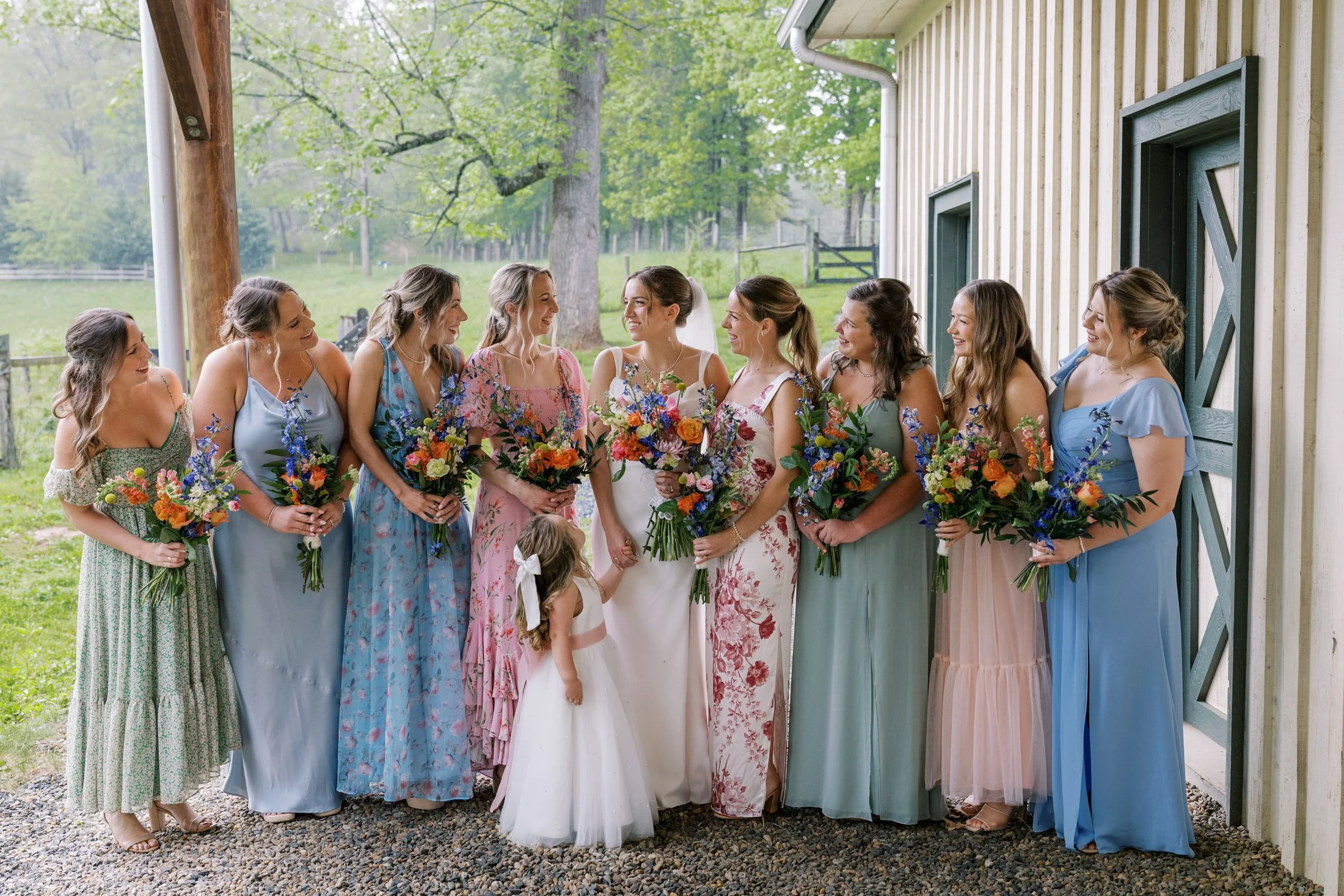 A group of women and two young girls at a wedding, standing outdoors near a rustic building with greenery in the background. The women are dressed in colorful dresses and holding bouquets of flowers, with the bride in the center wearing a white dress