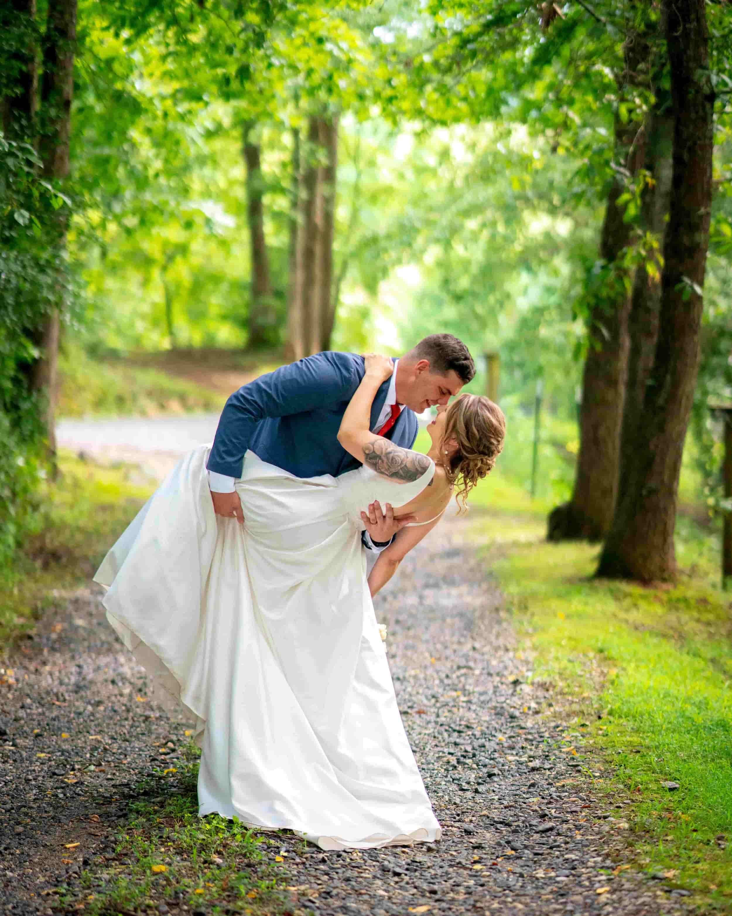 A bride and groom in a wooded outdoor setting, with the groom dipping the bride as they look at each other.