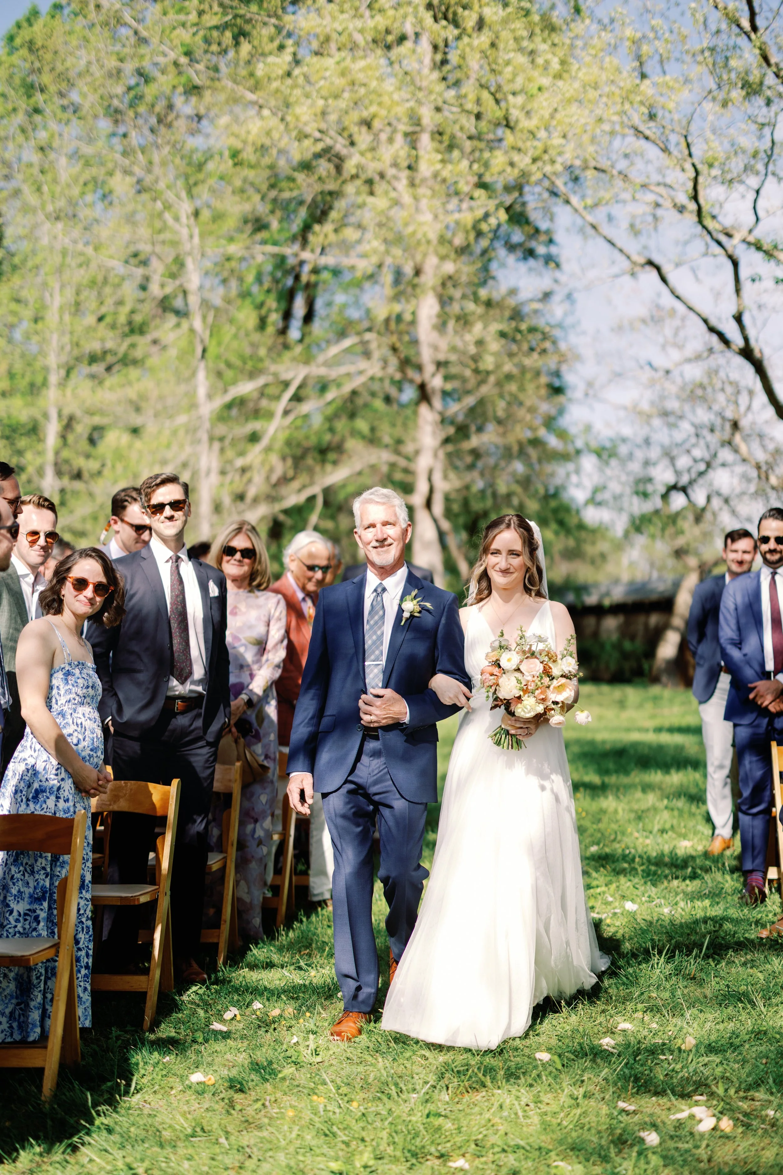A bride walking down the aisle arm-in-arm with an older man, possibly her father, on an outdoor wedding ceremony with guests seated on each side. She holds a bouquet of flowers and is wearing a white wedding dress. The setting is sunny with green trees and grass.