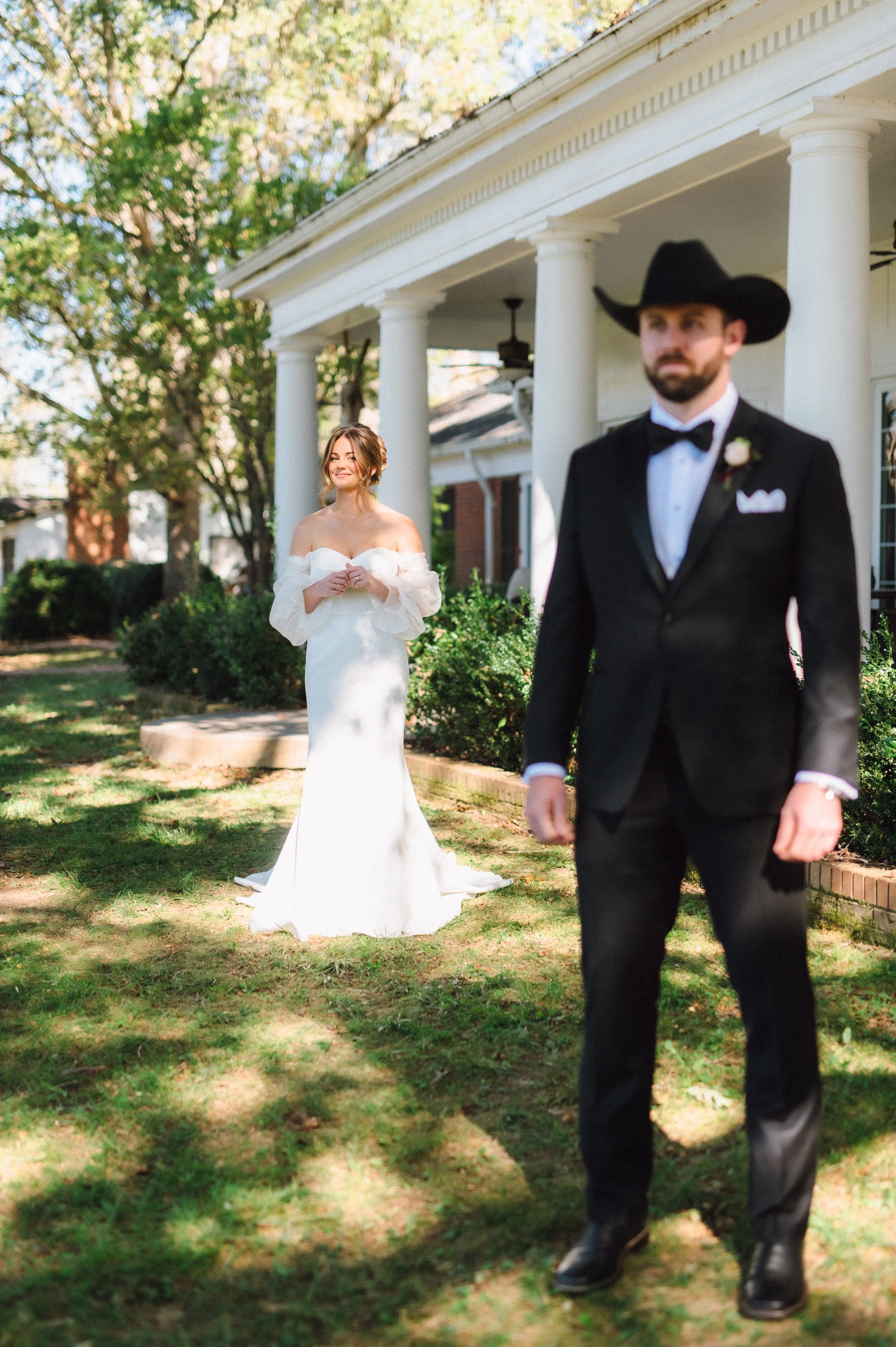 A bride in a white wedding dress with off-the-shoulder sleeves, standing on a grassy area with her hands clasped, smiling at a groom in a black tuxedo and cowboy hat, outside a white house with columns and trees.