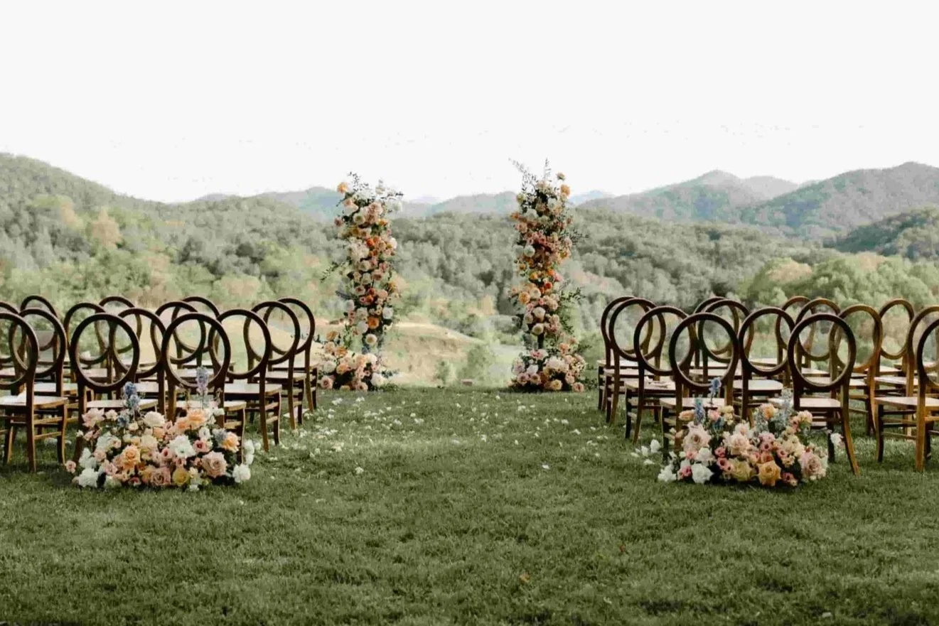Outdoor wedding setup with wooden chairs arranged on green grass, decorated with large floral arrangements and two tall floral archways, overlooking a scenic mountain landscape.