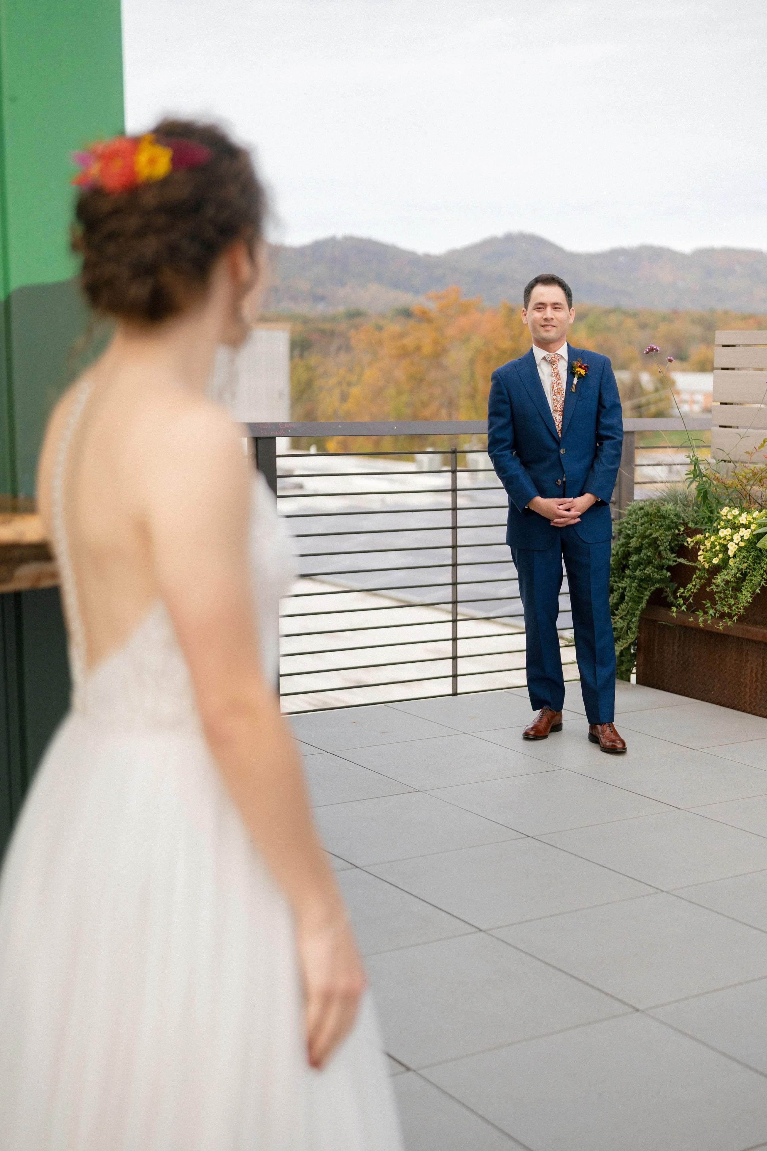 A bride in a white gown stands on a rooftop terrace, looking at a man in a blue suit standing in the distance with autumn foliage and mountains in the background.