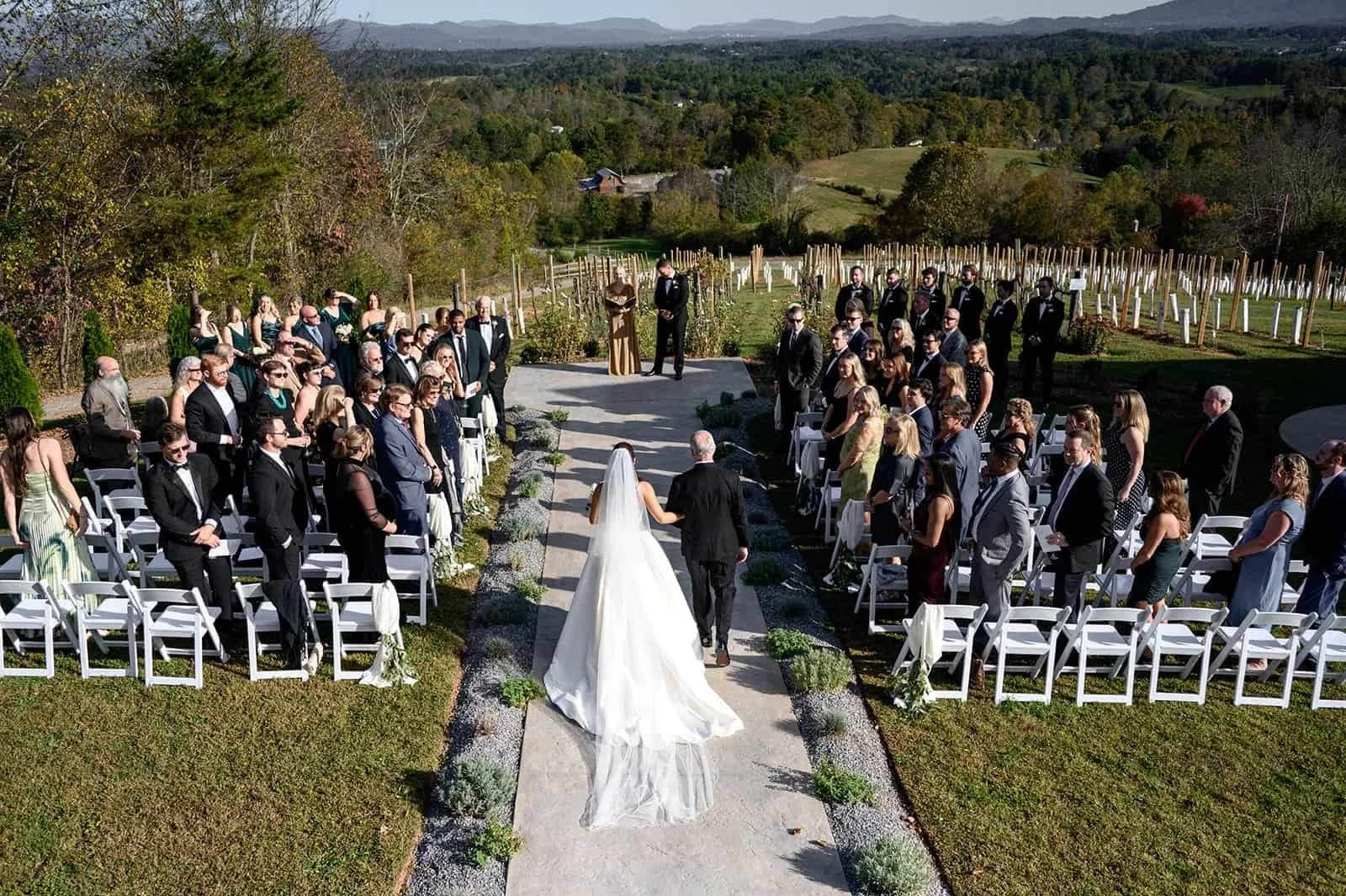 A wedding ceremony outdoors with a bride and officiant walking down the aisle lined with white chairs and small plants, surrounded by guests and scenic landscape with hills and trees.