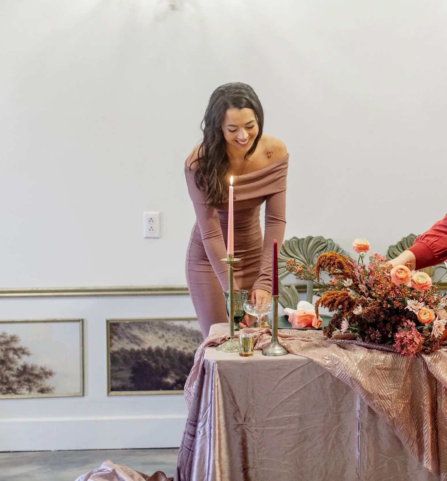 A woman in a mauve dress is smiling while leaning over a table with pink and gold decorations, candles, and a floral arrangement.
