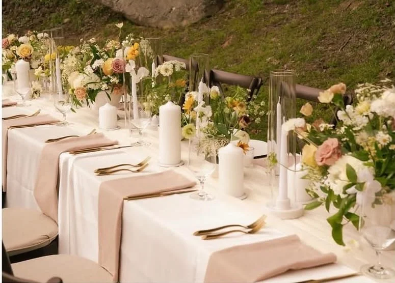 Elegant outdoor dining table set with white tablecloth, pink napkins, gold utensils, tall glass vases with white and pink flowers, white candles, and candles in cylindrical holders, surrounded by a grassy area.