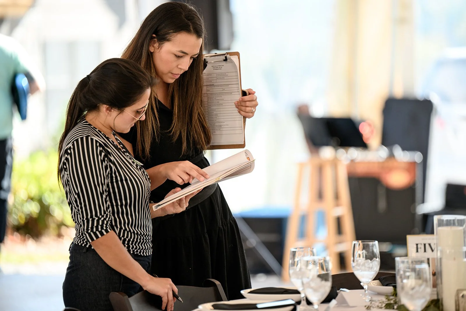 Two women standing at a restaurant table, reviewing a menu together, with glasses and table settings visible in the foreground.