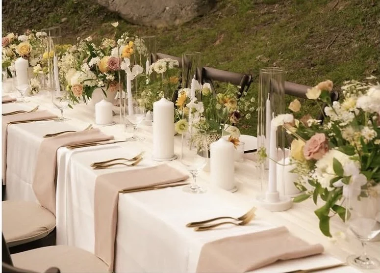 Elegant outdoor banquet table with white tablecloth, pink napkins, gold utensils, white candles, and floral centerpieces with pink, white, and yellow flowers.